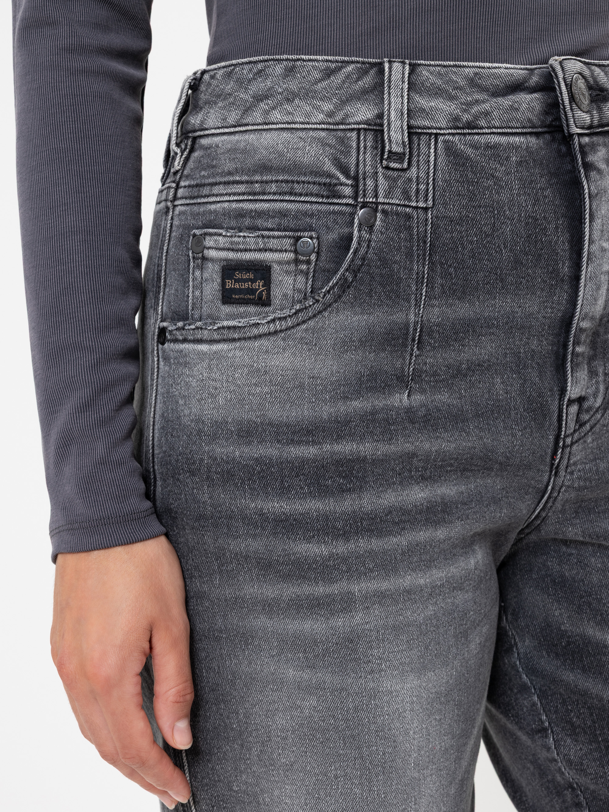 Eine Person, die eine verblichene schwarze Jeans mit hoher Taille und ein langärmeliges graues geripptes Oberteil trägt, wobei sie ihre linke Hand an der Seite ruhen lässt. Die Jeans hat sichtbare Nähte und ein kleines Markenetikett über der Vorderta
