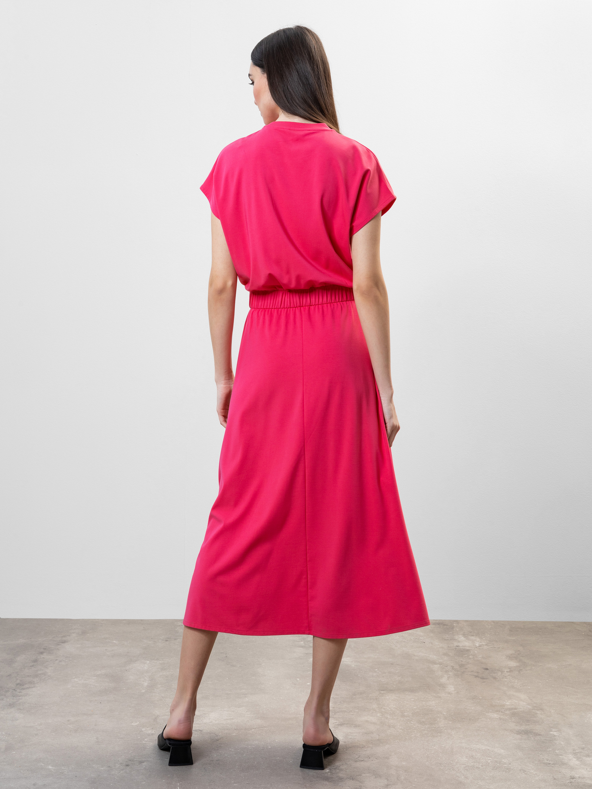 Eine Frau mit langen braunen Haaren steht in einem hellrosa, kurzärmeligen, halblangen Kleid mit elastischem Bund und schwarzen Sandaletten mit Absatz auf einem glatten Betonboden mit hellgrauem Hintergrund.