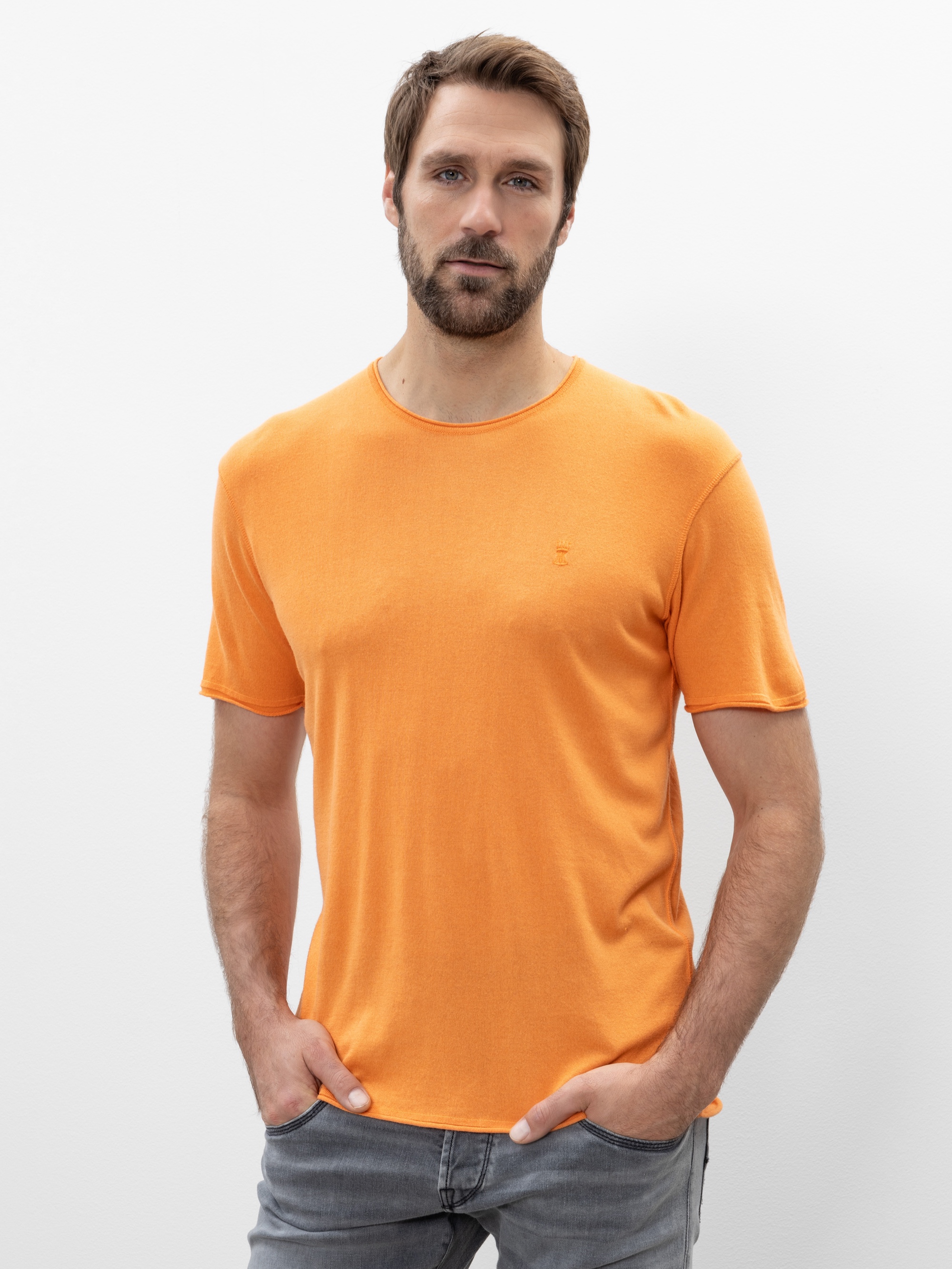 Vor einem schlichten weißen Hintergrund steht ein Mann mit Bart, der ein orangefarbenes kurzärmeliges T-Shirt und graue Jeans trägt und die Hände in den Taschen hat.