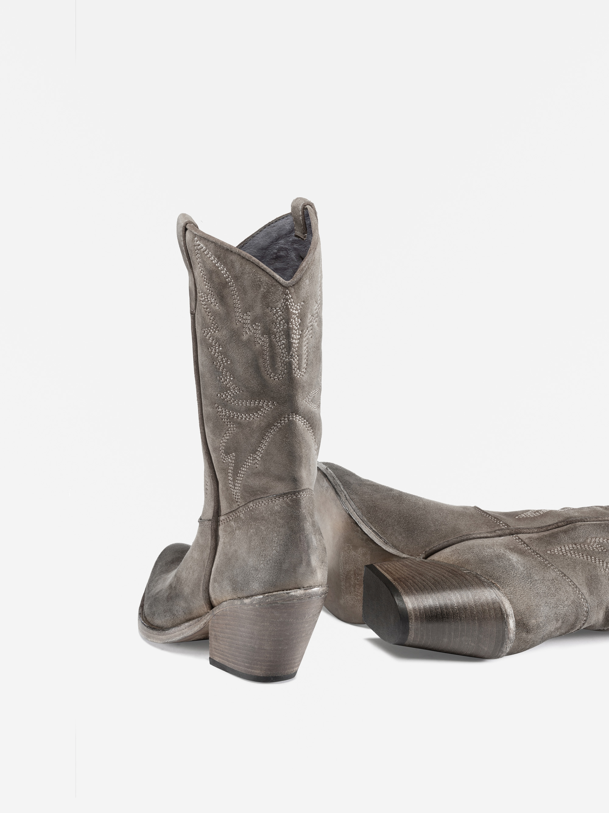 Ein Paar hellgraue Cowboystiefel aus Wildleder mit gestickten Mustern, spitzen Zehen und gestapelten Holzabsätzen sind vor einem weißen Hintergrund abgebildet. Ein Stiefel steht aufrecht, während der andere auf der Seite liegt.