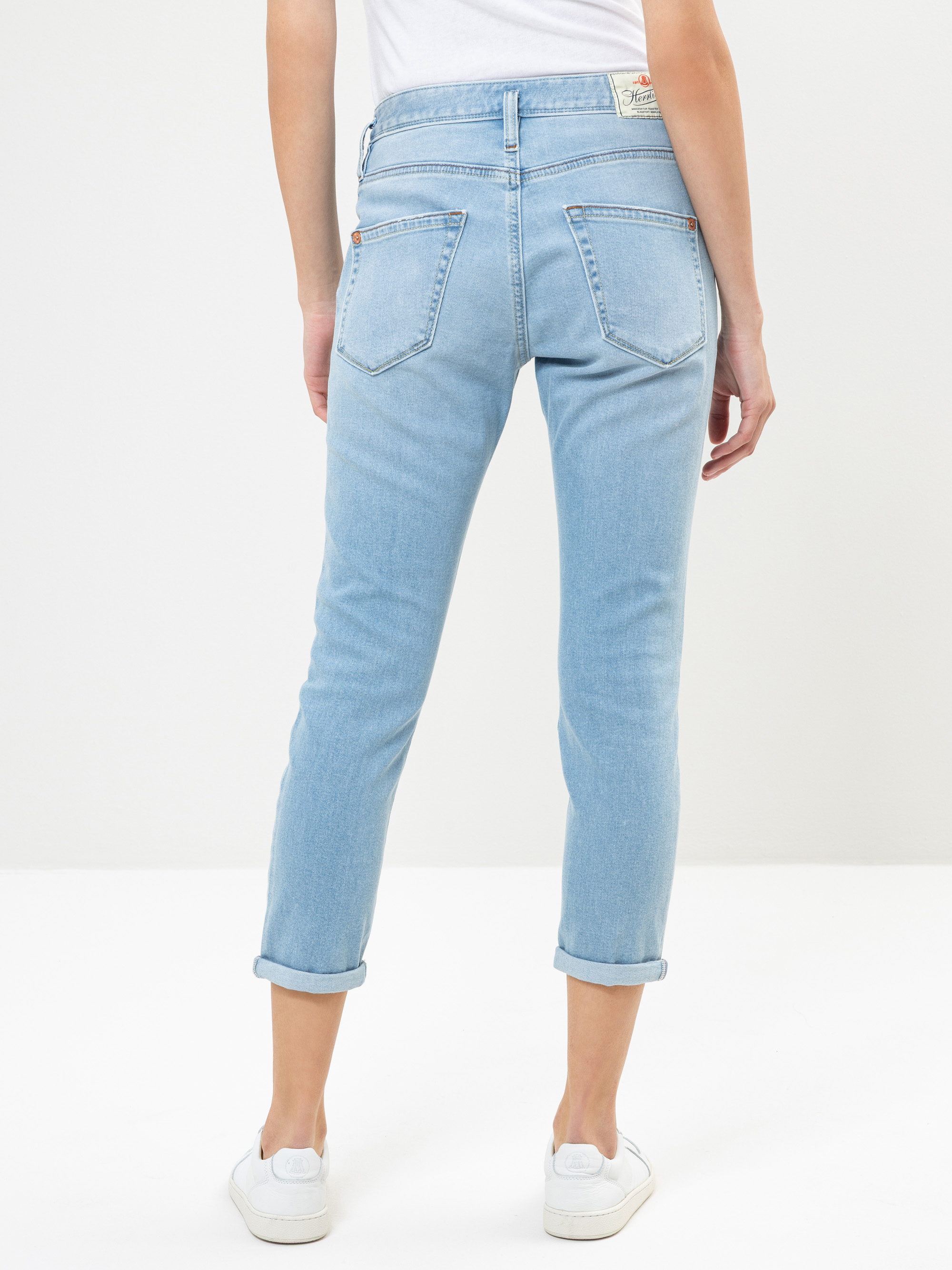 Eine Person in hellblauen Jeans mit Bündchen und weißen Turnschuhen steht mit dem Gesicht zur Kamera vor einem schlichten weißen Hintergrund.