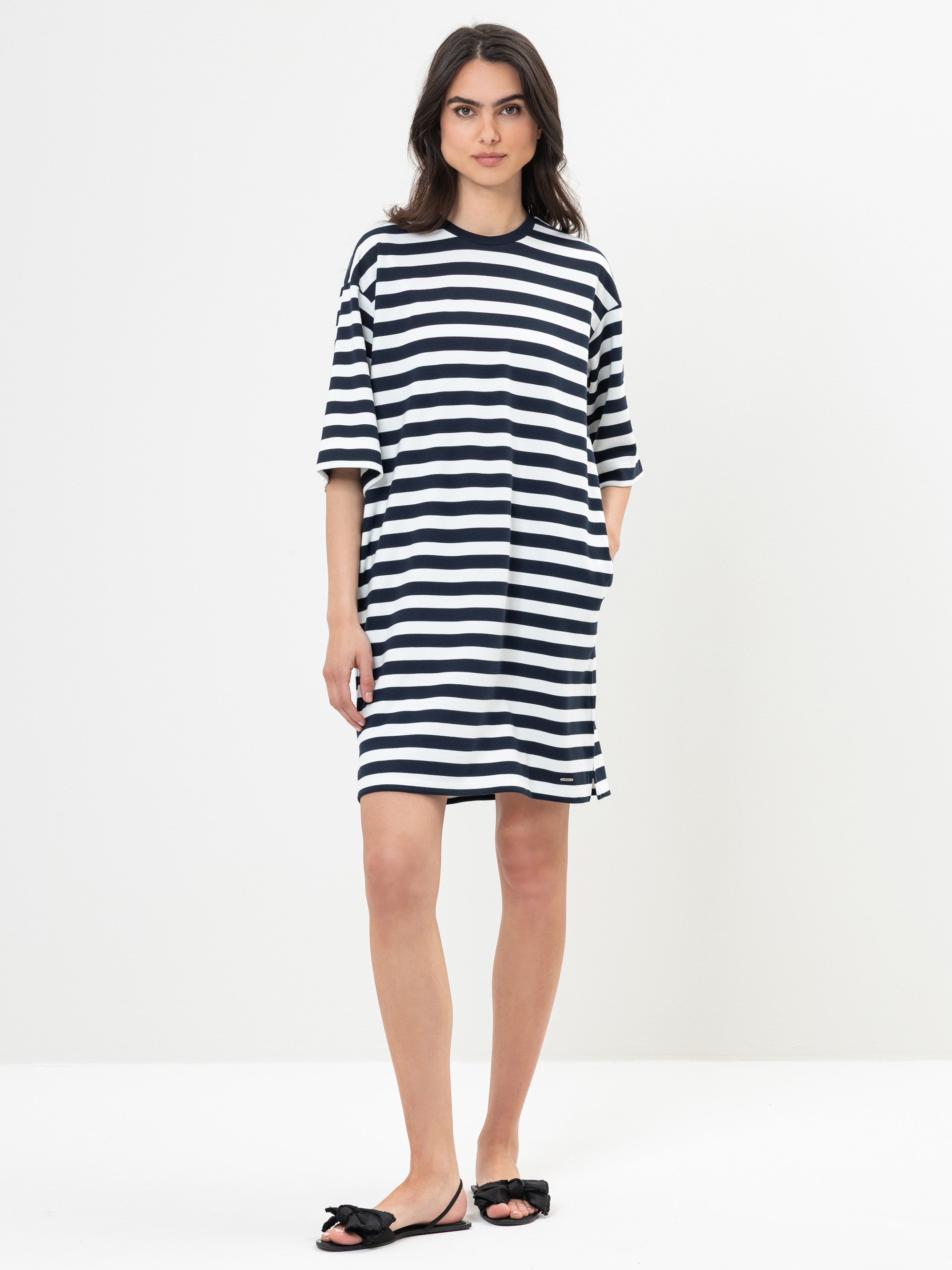 Eine Frau steht vor einem schlichten weißen Hintergrund und trägt ein locker sitzendes, knielanges Kleid mit schwarzen und weißen Querstreifen. Sie hat langes dunkles Haar und trägt schwarze Gleitsandalen mit Schleifen.