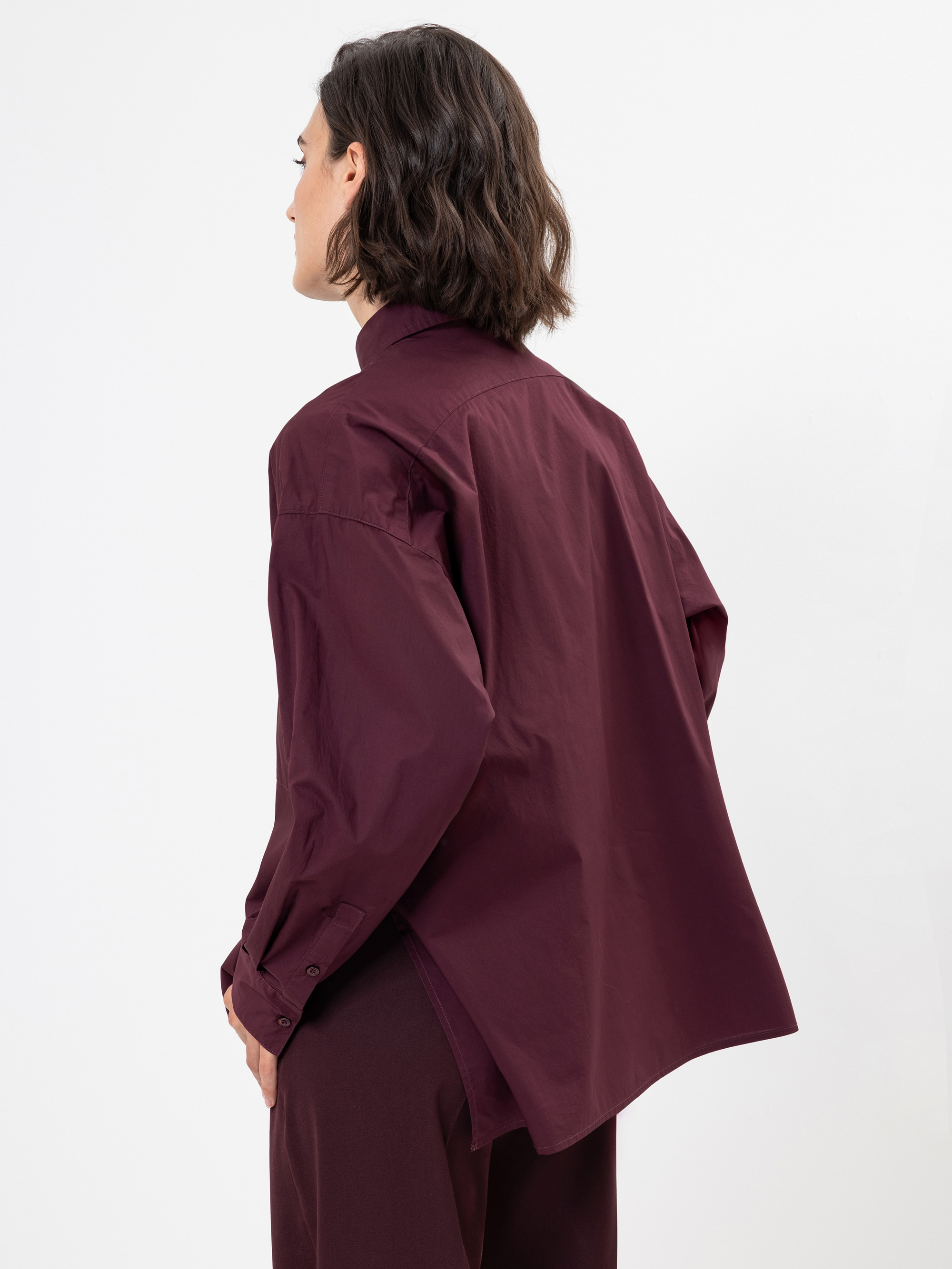 Eine Person mit kurzen braunen Haaren, die ein locker sitzendes kastanienbraunes Button-up-Hemd und eine passende Hose trägt, steht mit dem Rücken zur Kamera vor einem schlichten weißen Hintergrund.