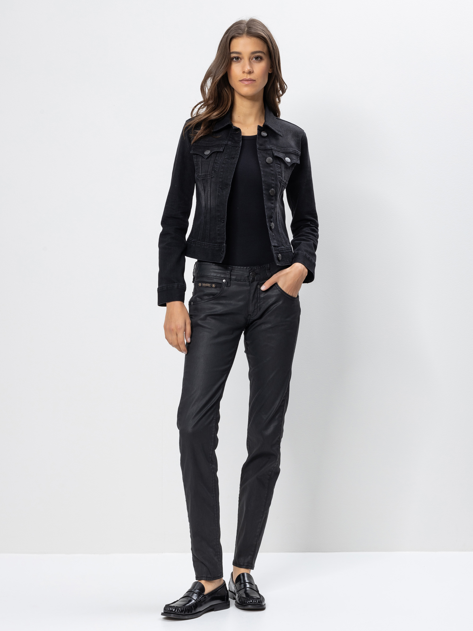 Eine Frau steht vor einem weißen Hintergrund, trägt eine schwarze Jeansjacke, ein schwarzes Oberteil, eine schwarze Hose und schwarze Halbschuhe, eine Hand in der Tasche und einen neutralen Gesichtsausdruck.