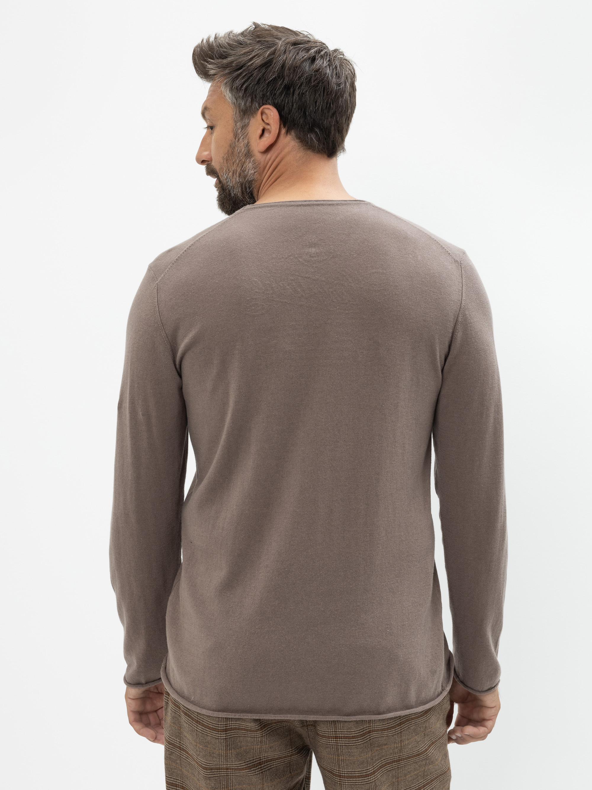 Ein Mann mit kurzen dunklen Haaren und Bart steht mit dem Rücken zur Kamera, trägt ein langärmeliges taupefarbenes Hemd und eine karierte Hose vor einem schlichten weißen Hintergrund.