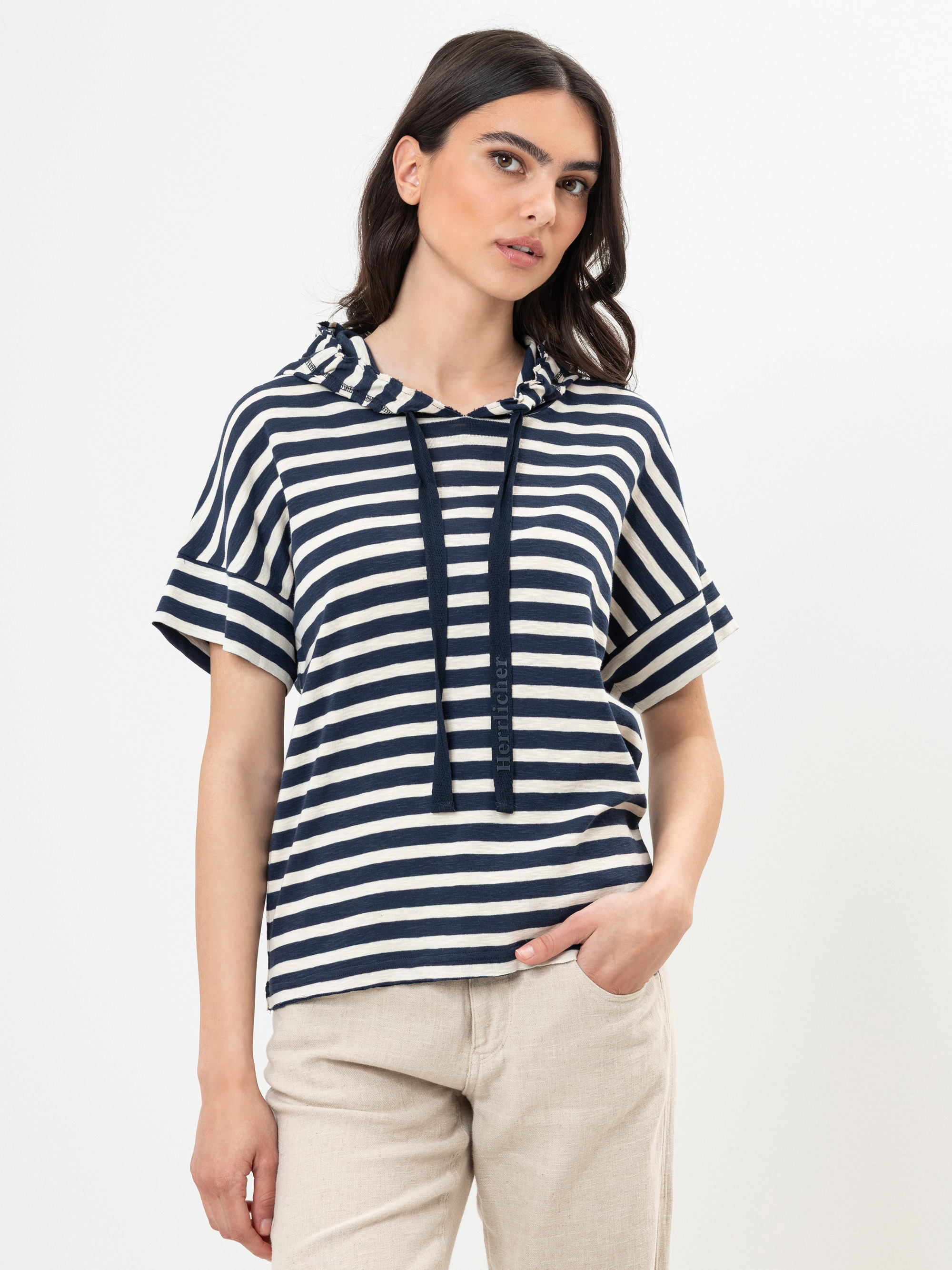 Eine Frau, die ein marineblau-weiß gestreiftes, kurzärmeliges Kapuzenoberteil mit Kordelzug und eine hellbeige Hose trägt, steht vor einem schlichten weißen Hintergrund.