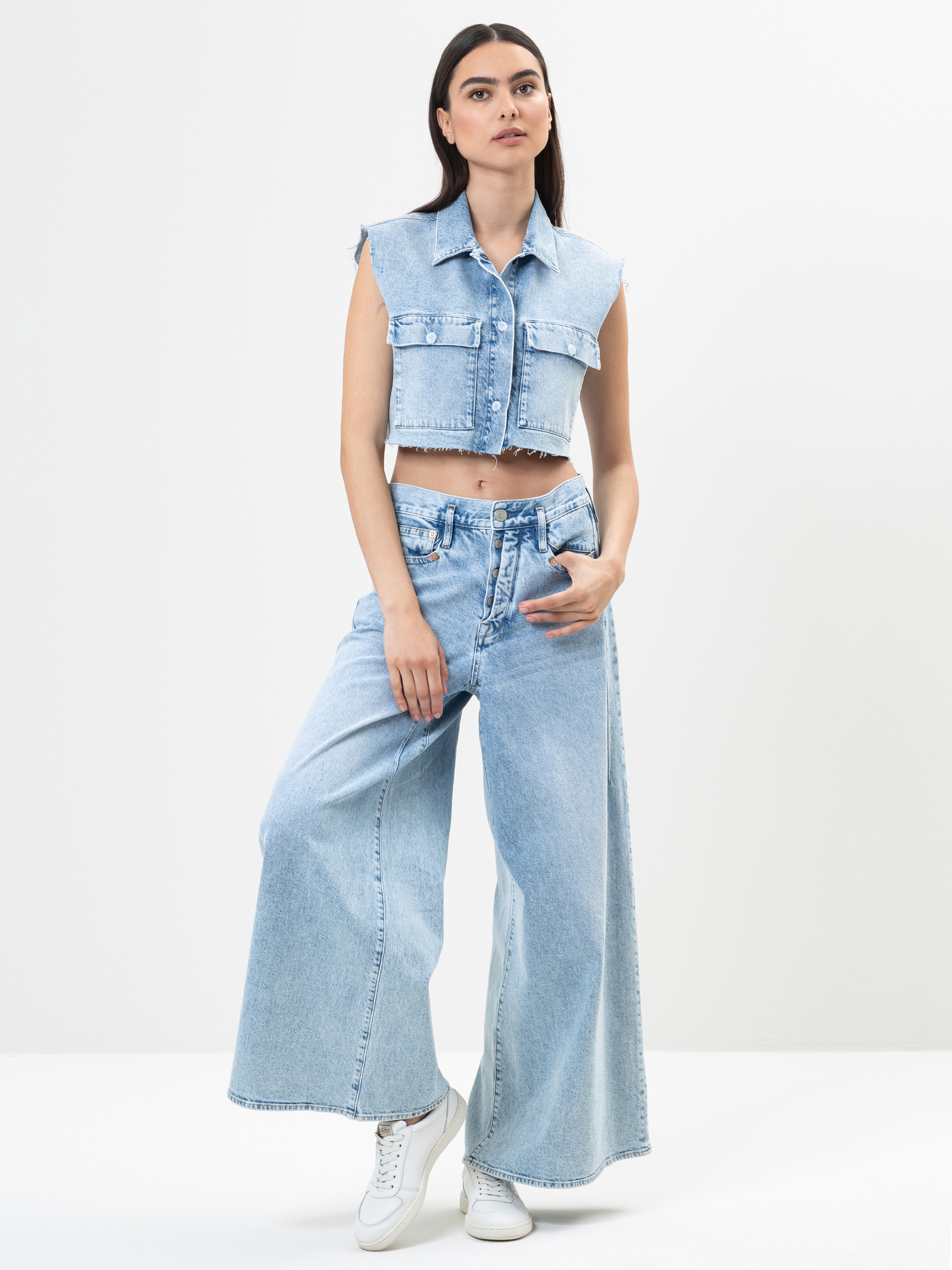 Eine Frau steht vor einem schlichten weißen Hintergrund und trägt eine ärmellose Jeansweste, weite Jeans mit hoher Taille und weiße Turnschuhe. Sie hat langes dunkles Haar und einen neutralen Gesichtsausdruck.