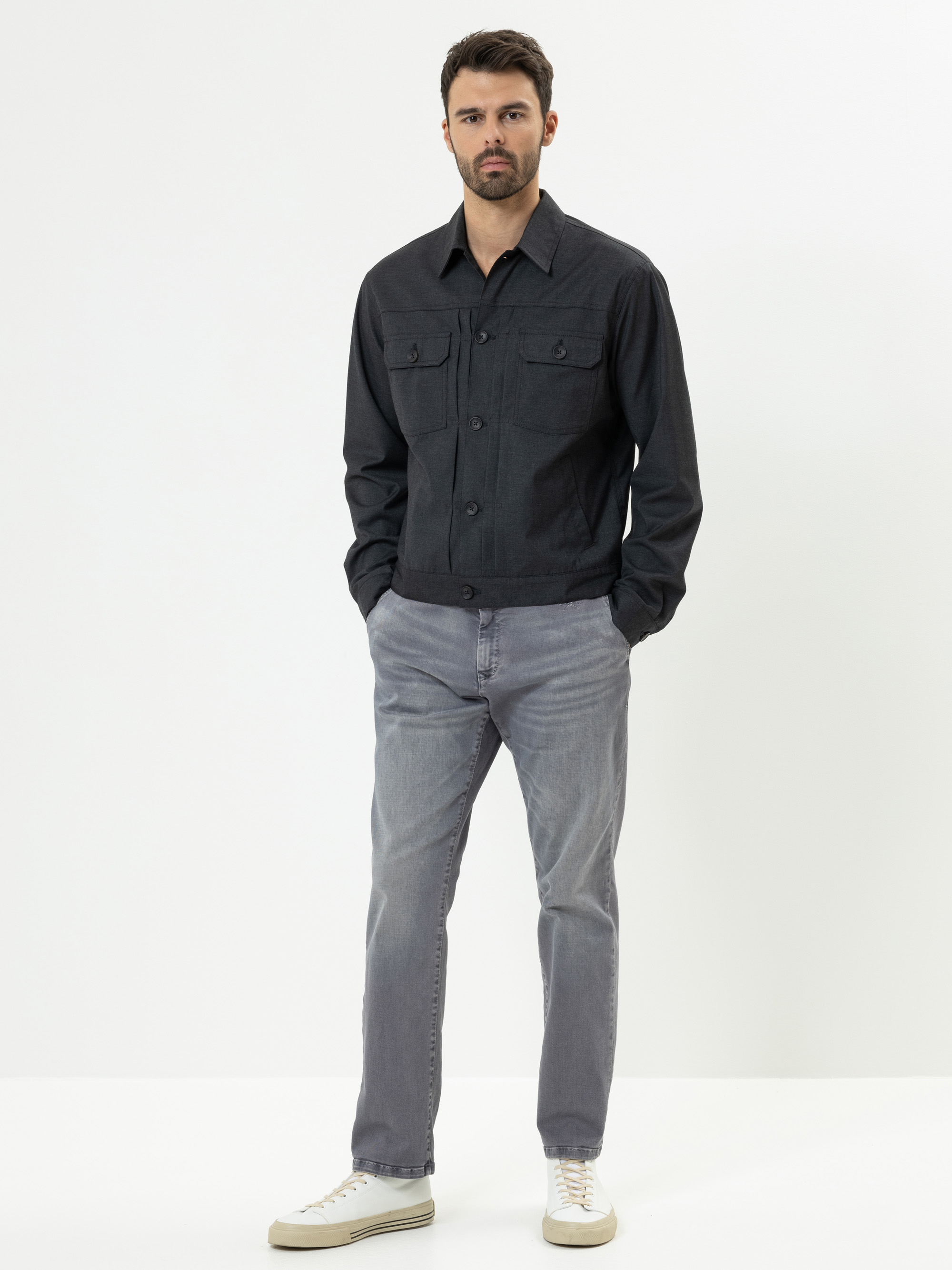 Ein Mann steht vor einem schlichten weißen Hintergrund und trägt eine dunkle Button-up-Jacke, hellgraue Jeans und weiße Turnschuhe. Er hat kurze Haare und einen Bart, steht mit den Händen in den Taschen und einem neutralen Gesichtsausdruck.
