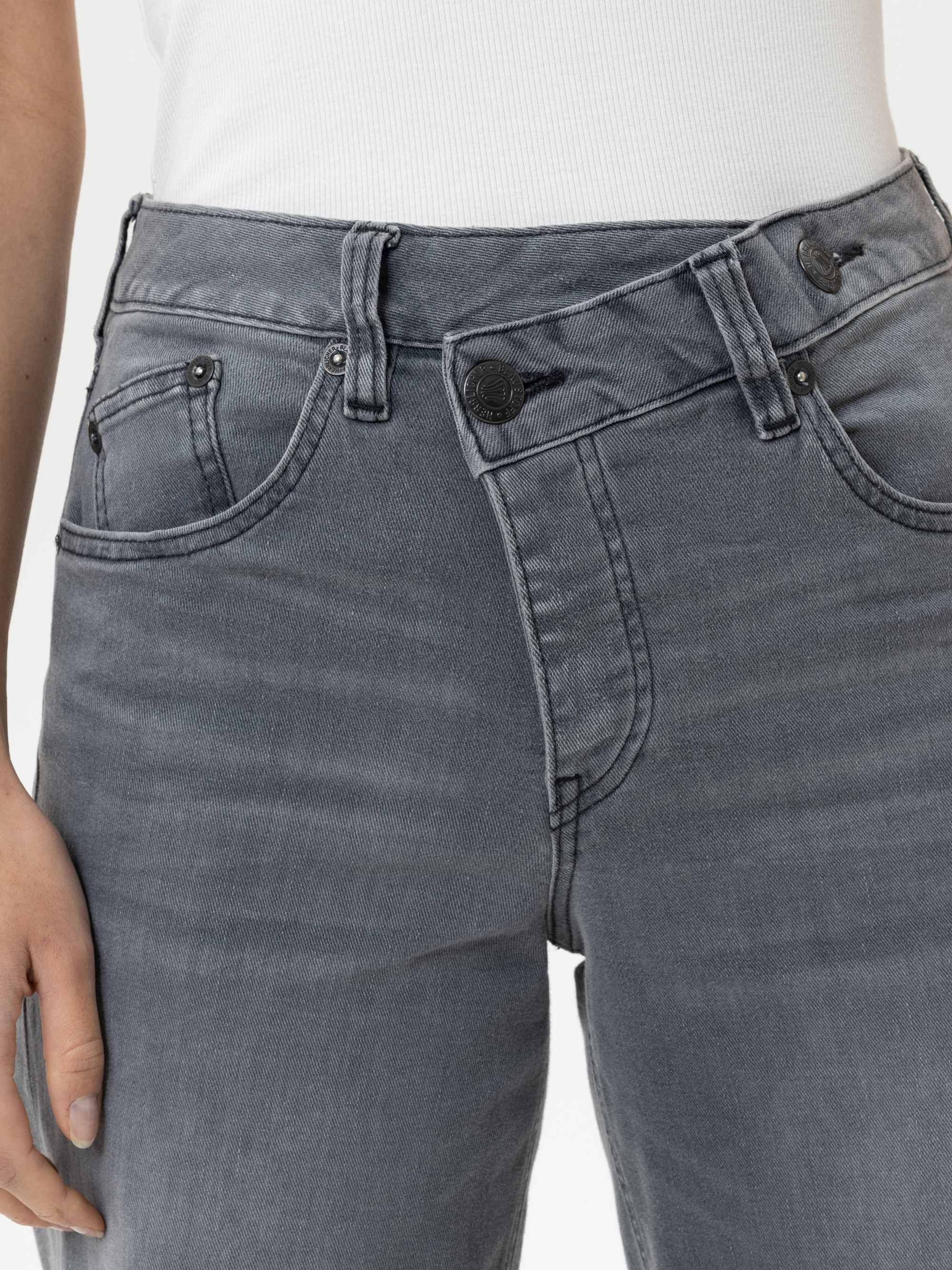 Eine Person trägt eine graue Jeans mit hoher Taille und einem einzigartigen diagonalen Doppel-Bund-Design, gepaart mit einem weißen Oberteil. Die linke Hand der Person ist an ihrer Seite zu sehen.