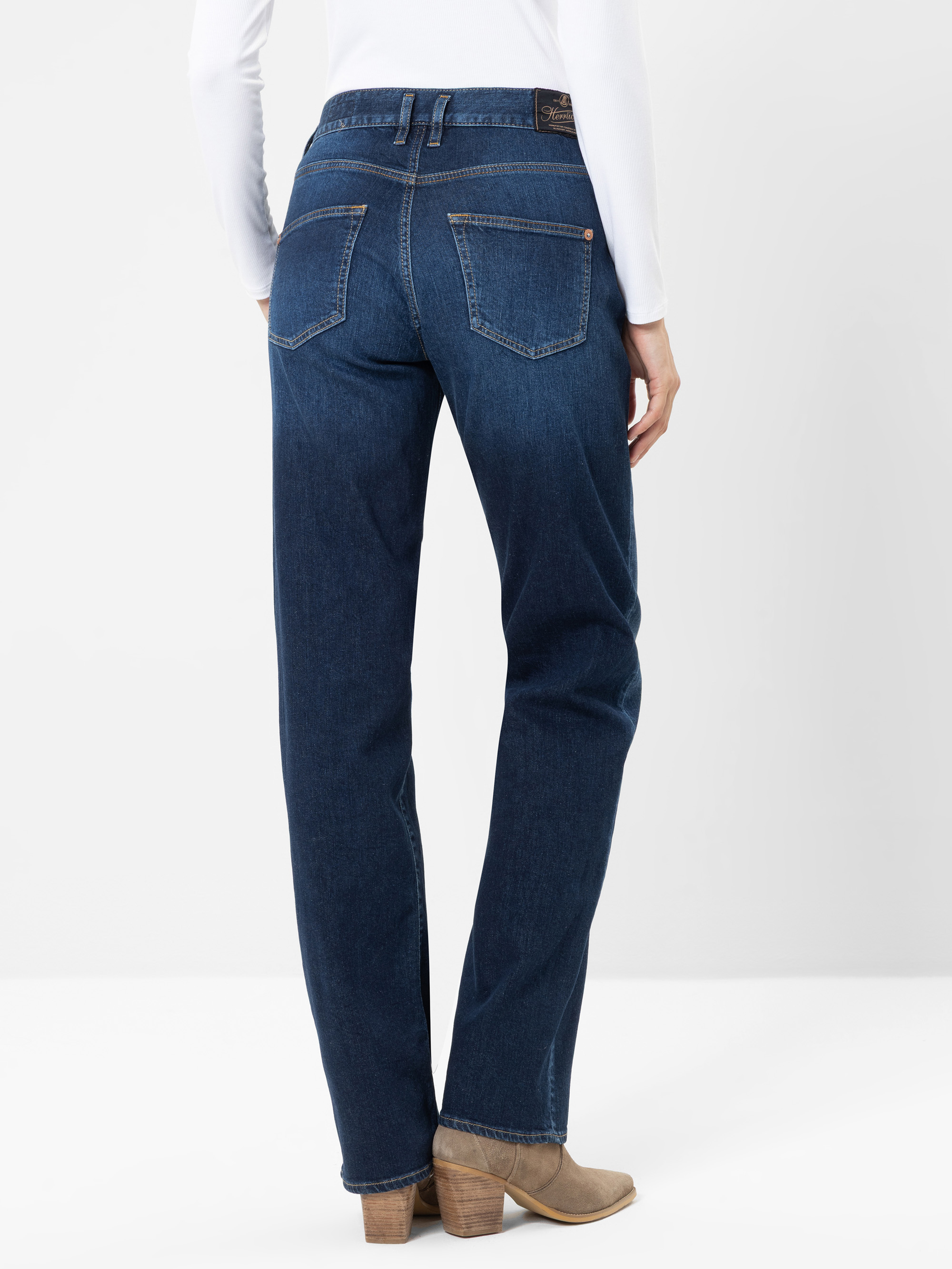 Eine Person, die dunkelblaue Jeans, ein weißes Langarm-Oberteil und hellbraune Stiefel mit Absatz trägt, von hinten vor einem schlichten weißen Hintergrund abgebildet.