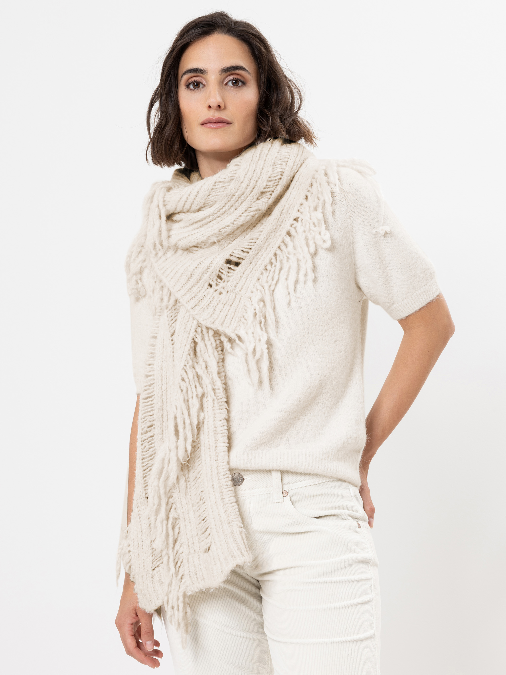 Eine Frau, die einen cremefarbenen, kurzärmeligen Pullover, eine dazu passende Hose und einen dicken, fransenbesetzten Strickschal trägt, steht vor einem schlichten weißen Hintergrund und blickt selbstbewusst in die Kamera.
