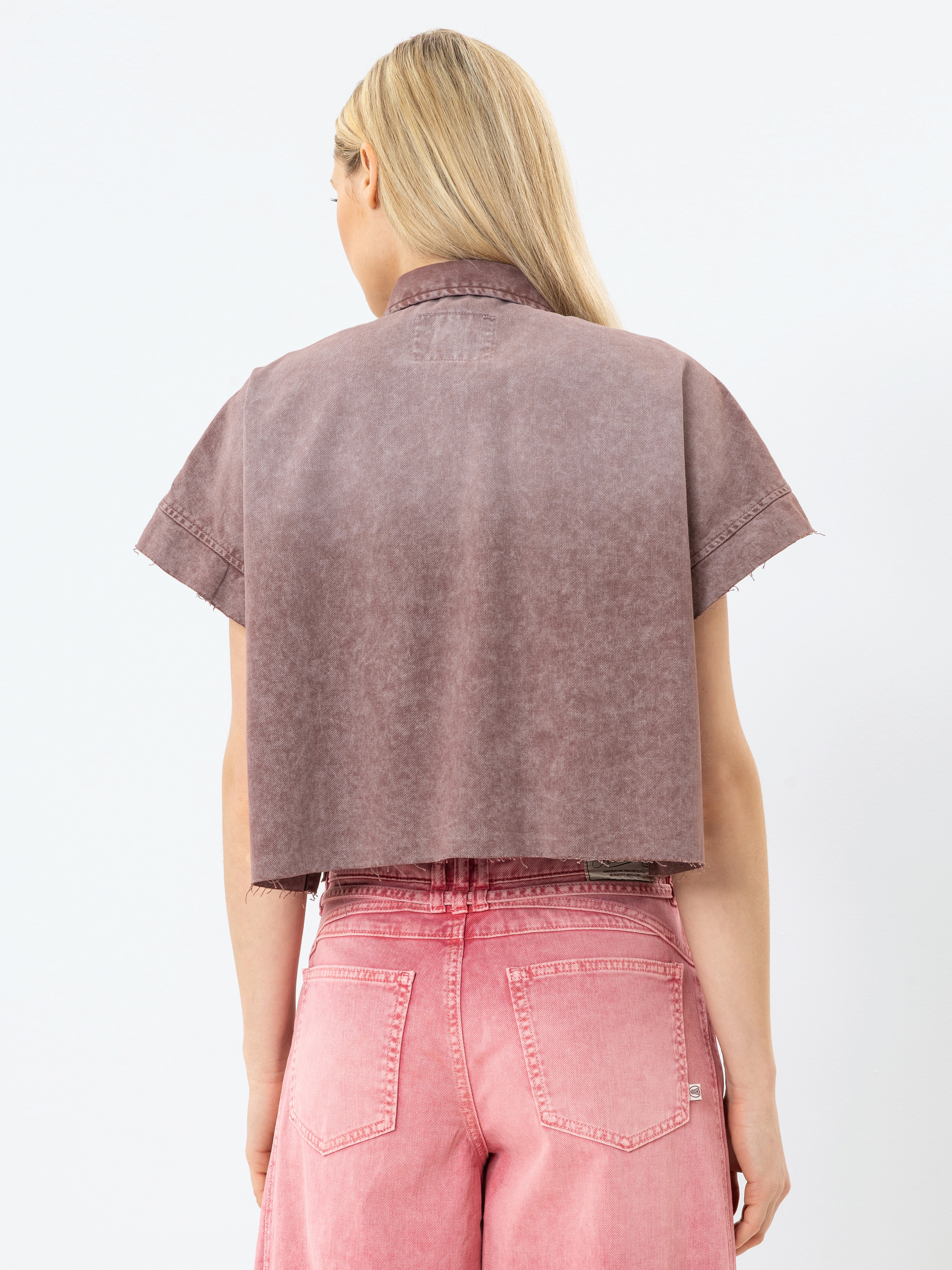 Eine Person mit langen blonden Haaren trägt eine verblasste lila Kurzarm-Jacke und hochgekrempelte rosa Jeans und steht mit dem Rücken zur Kamera vor einem schlichten weißen Hintergrund.