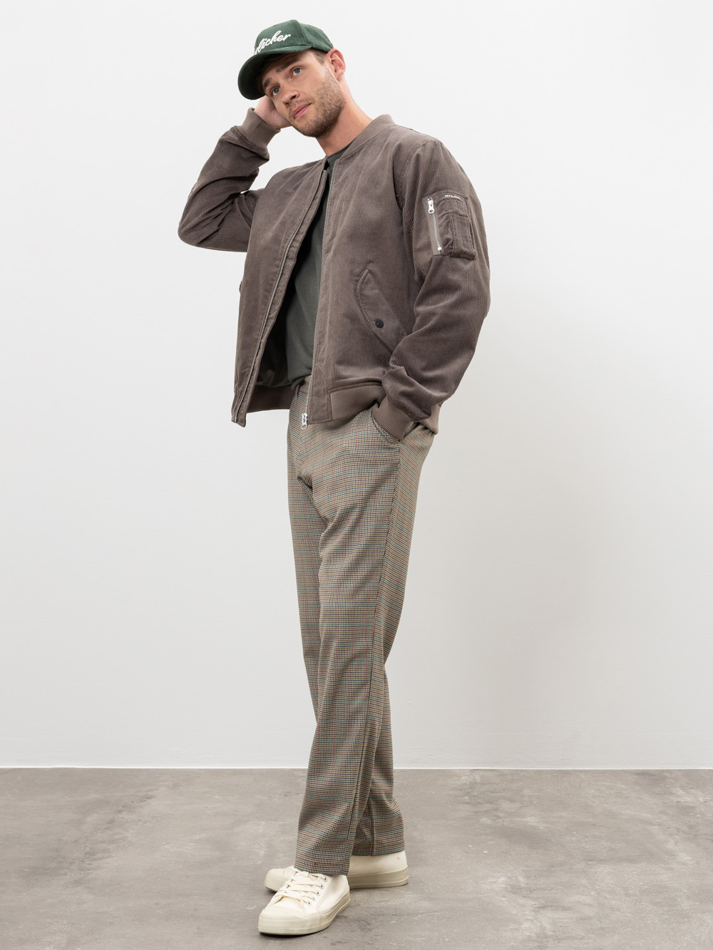 Ein Mann steht in entspannter Pose mit grüner Mütze, brauner Bomberjacke, dunklem Hemd, karierter Hose und cremefarbenen Turnschuhen vor einer schlichten weißen Wand und einem grauen Boden.