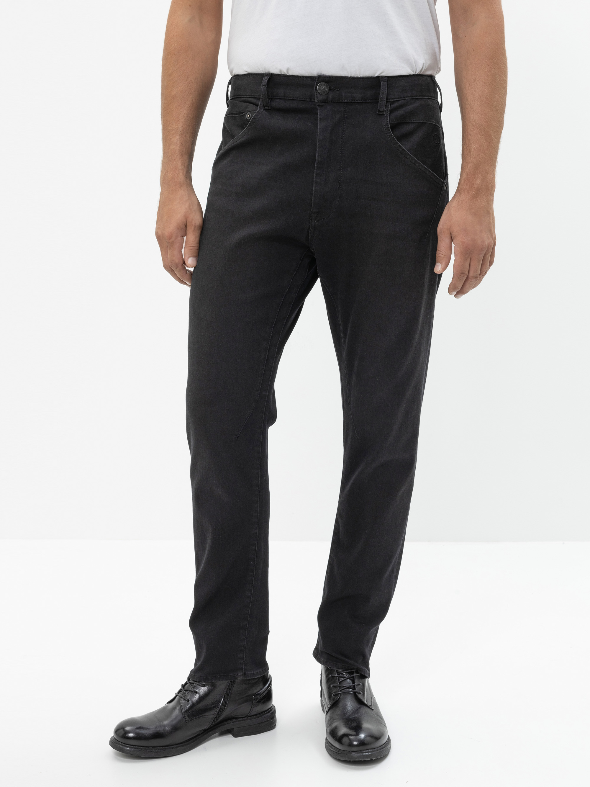 Ein Mann, der ein weißes T-Shirt, schwarze Jeans und schwarze Schuhe trägt, steht vor einem schlichten weißen Hintergrund und hat die Arme locker an den Seiten.