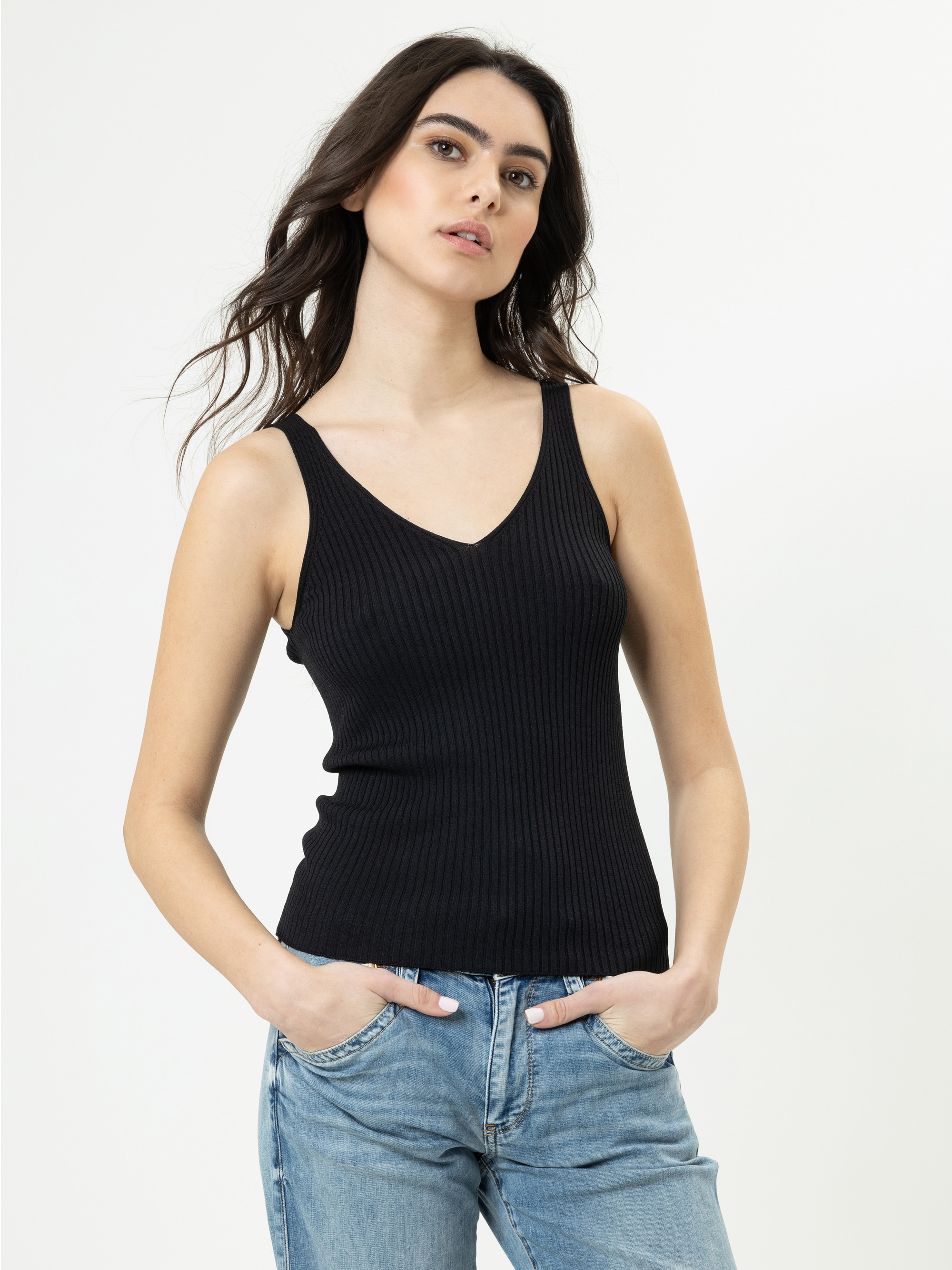 Eine Frau mit langen dunklen Haaren trägt ein ärmelloses schwarzes Oberteil mit geripptem V-Ausschnitt und hellblaue Jeans und steht mit den Händen in den Taschen vor einem schlichten weißen Hintergrund.