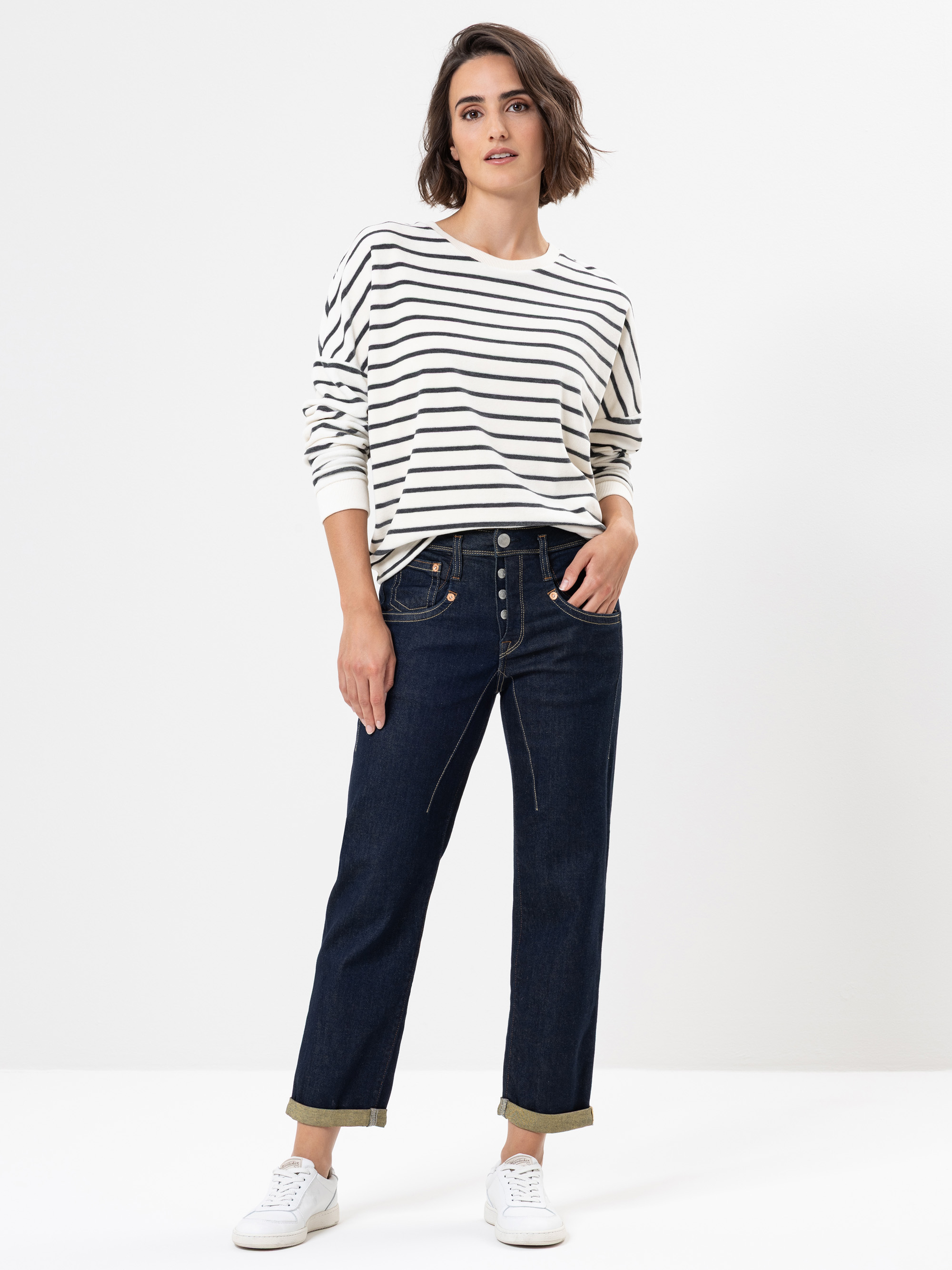 Eine Person, die ein weißes langärmeliges Hemd mit schwarzen Querstreifen, dunkle Jeans mit Bündchen und weiße Turnschuhe trägt, steht mit einer Hand in der Tasche vor einem schlichten weißen Hintergrund.