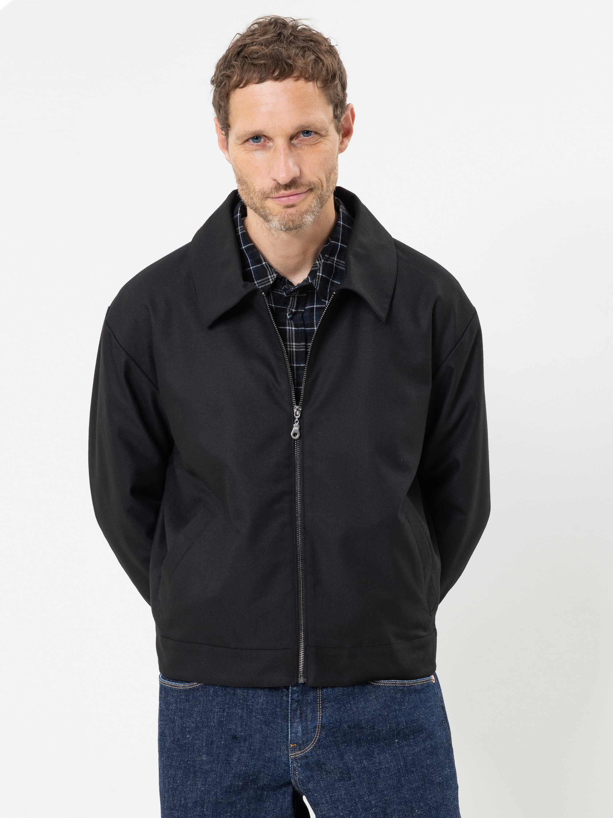 Ein Mann mit kurzen braunen Haaren und einem gepflegten Bart trägt eine schwarze Jacke mit Reißverschluss über einem karierten Hemd und blauen Jeans und steht mit hinter dem Rücken verschränkten Händen vor einem schlichten weißen Hintergrund.