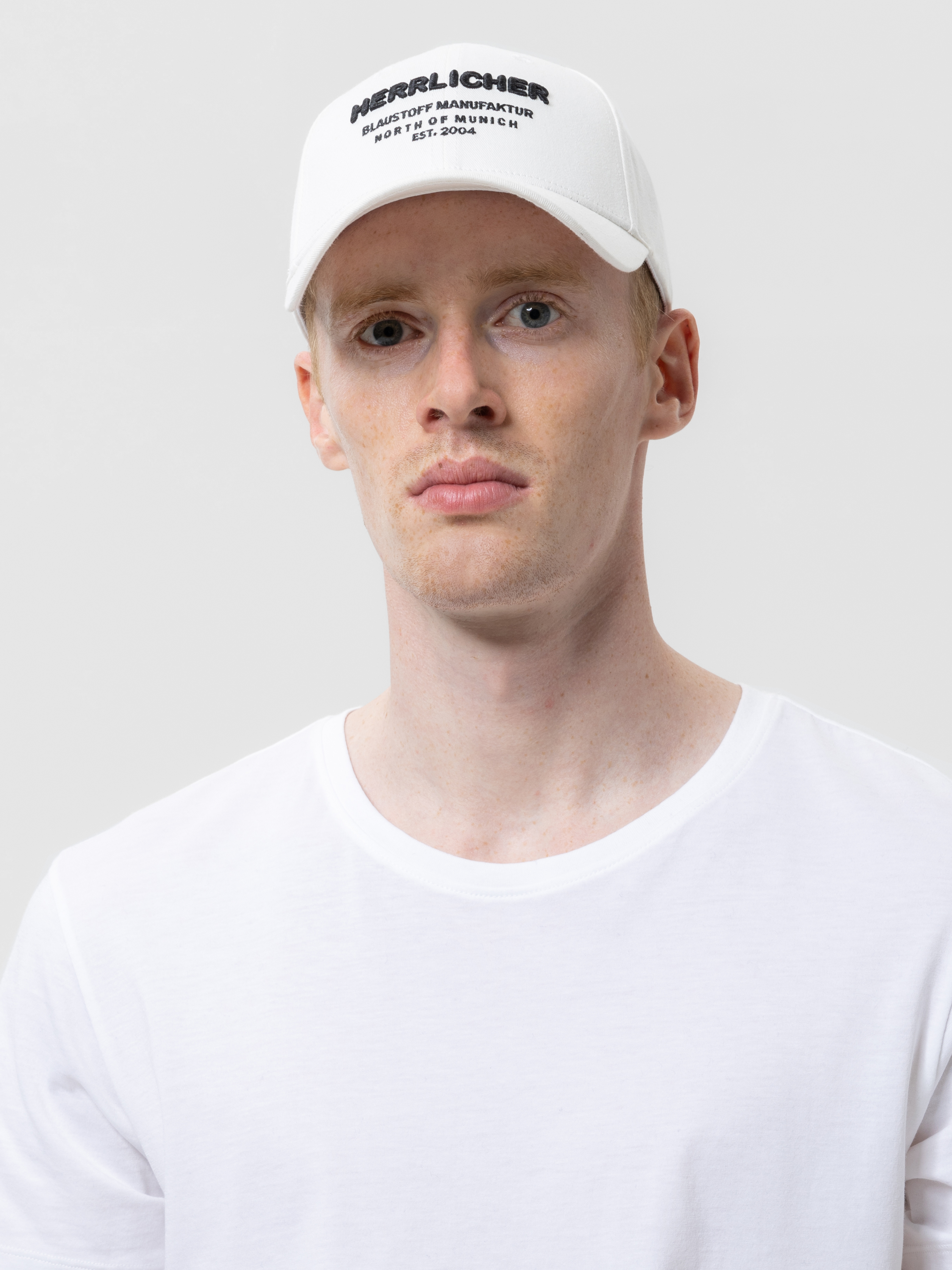 Eine Person, die ein schlichtes weißes T-Shirt und eine weiße Baseballkappe mit schwarzem Text auf der Vorderseite trägt, steht vor einem hellgrauen Hintergrund. Die Person hat einen neutralen Gesichtsausdruck.