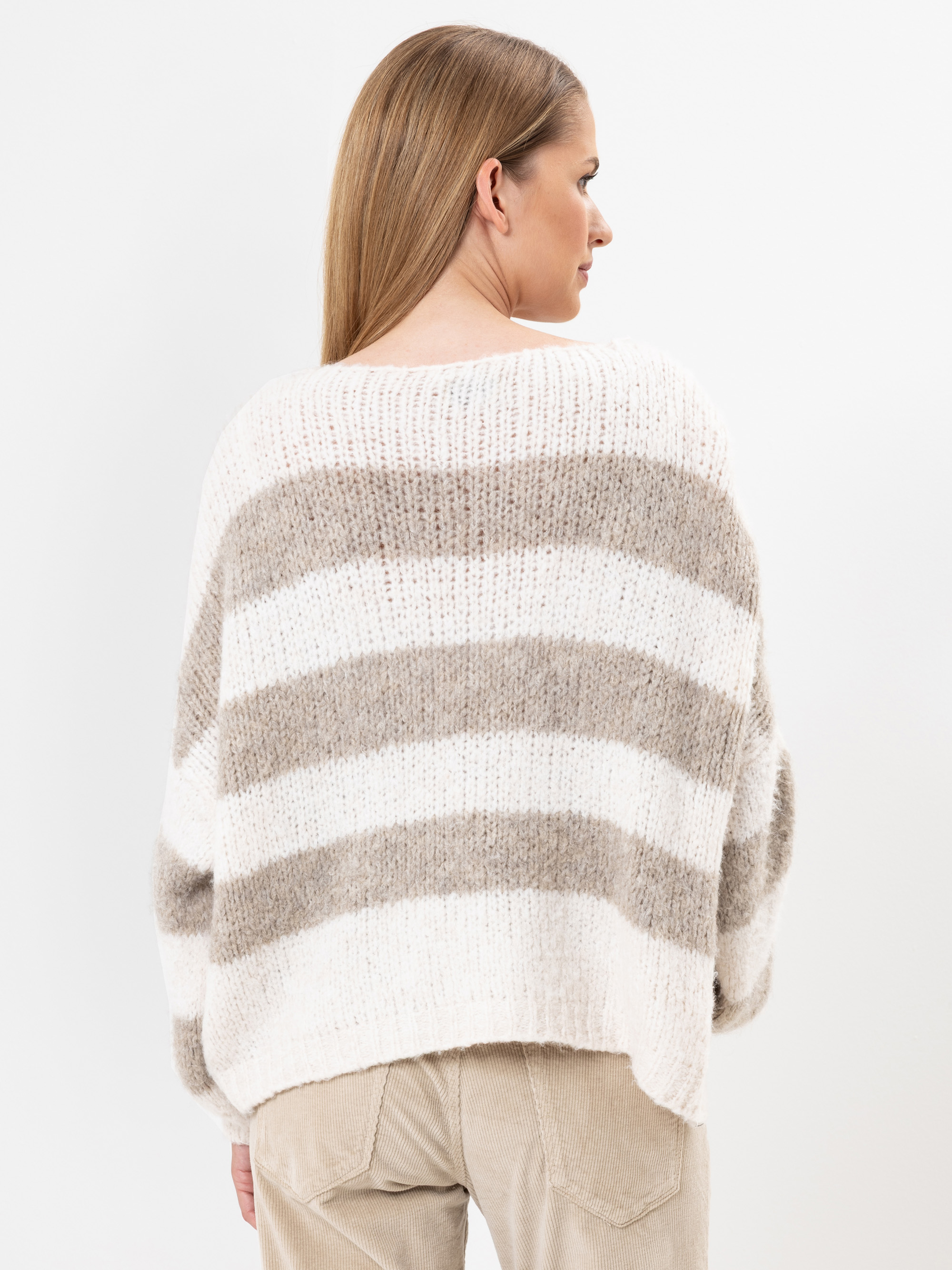 Eine Frau mit glattem, hellbraunem Haar trägt einen lockeren, dicken Strickpullover mit breiten horizontalen beigen und weißen Streifen und steht mit dem Rücken zur Kamera vor einem schlichten weißen Hintergrund.