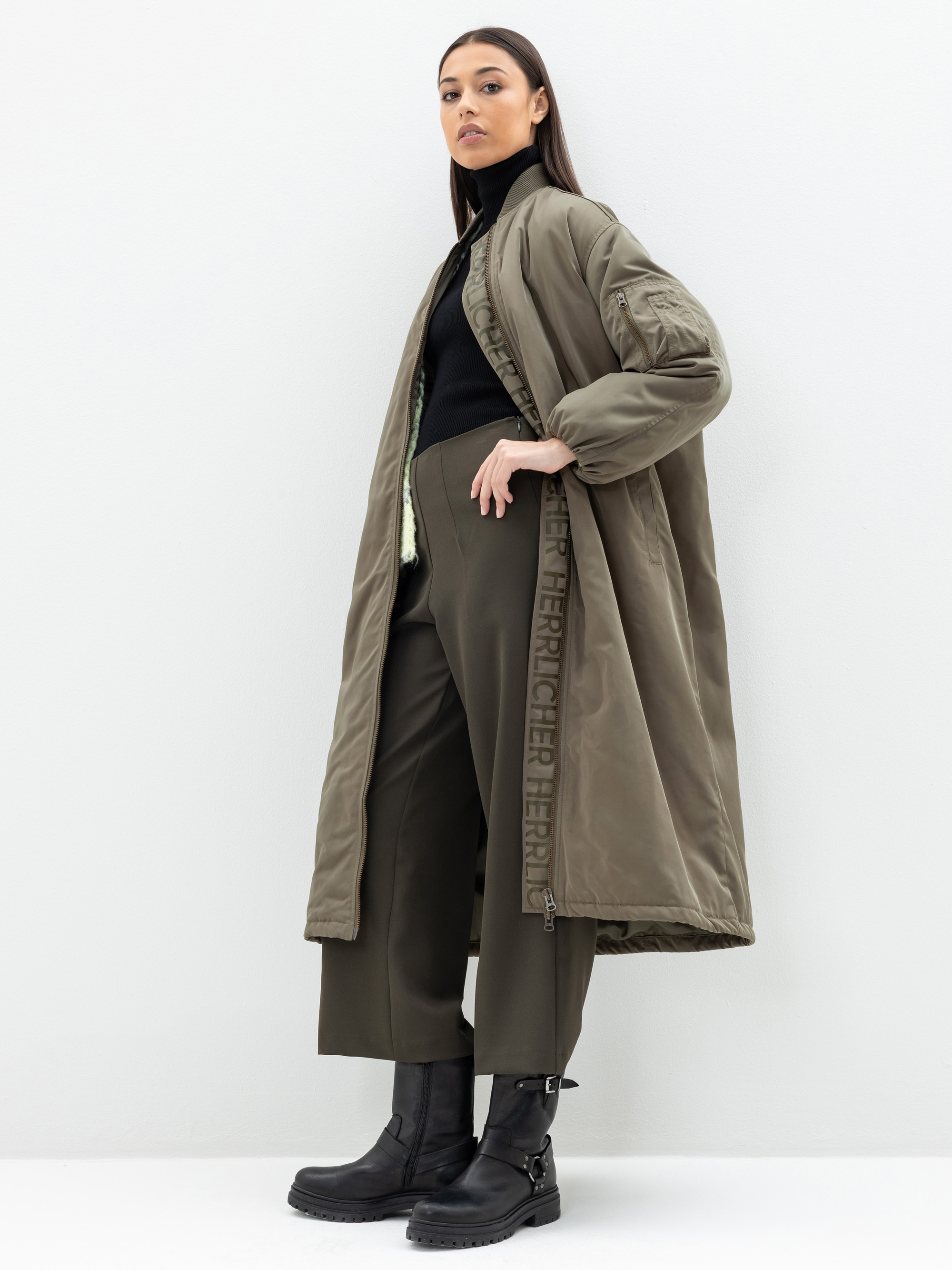 Eine Frau steht vor einem weißen Hintergrund und trägt einen olivgrünen, langen Steppmantel, eine dunkelgrüne Hose, einen schwarzen Rollkragenpullover und schwarze Stiefel. Sie blickt selbstbewusst in die Kamera und hat eine Hand in ihrer Tasche.