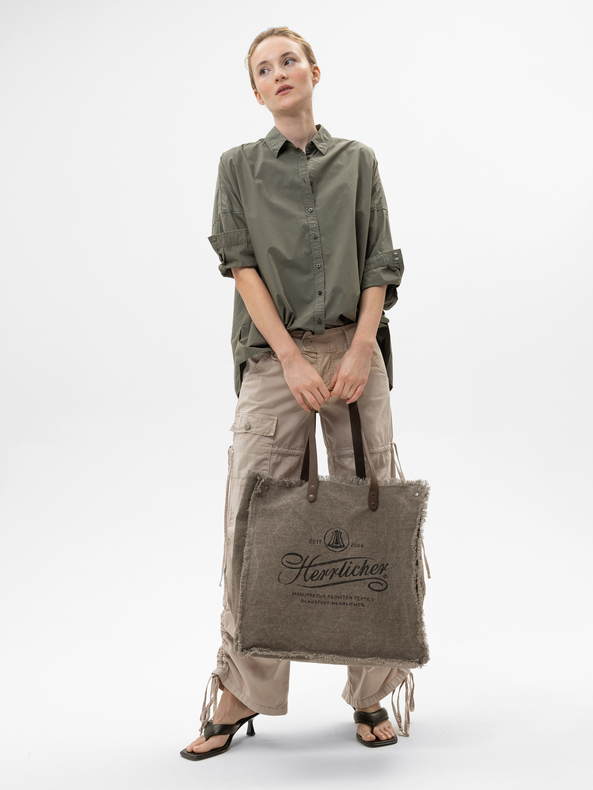Eine Frau, die ein lockeres olivgrünes Hemd und eine beigefarbene Cargohose trägt, steht vor einem weißen Hintergrund und hält eine große braune strukturierte Tragetasche mit Text darauf. Sie trägt schwarze, hochhackige Sandalen.
