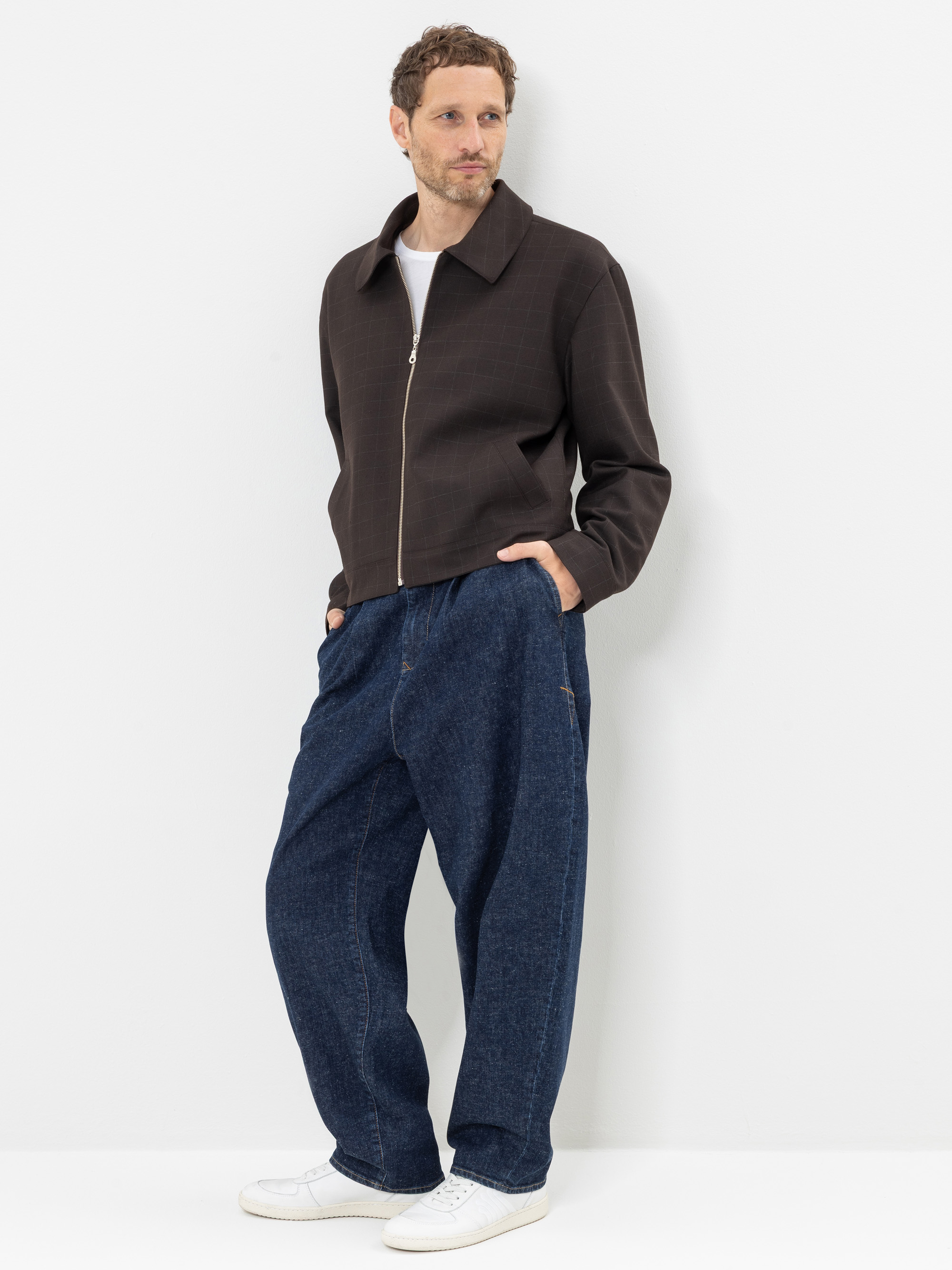 Ein Mann steht vor einer weißen Wand. Er trägt eine dunkle Jacke mit Reißverschluss, weite blaue Jeans, ein weißes T-Shirt und weiße Turnschuhe, hat die Hände in den Taschen und schaut leicht zur Seite.