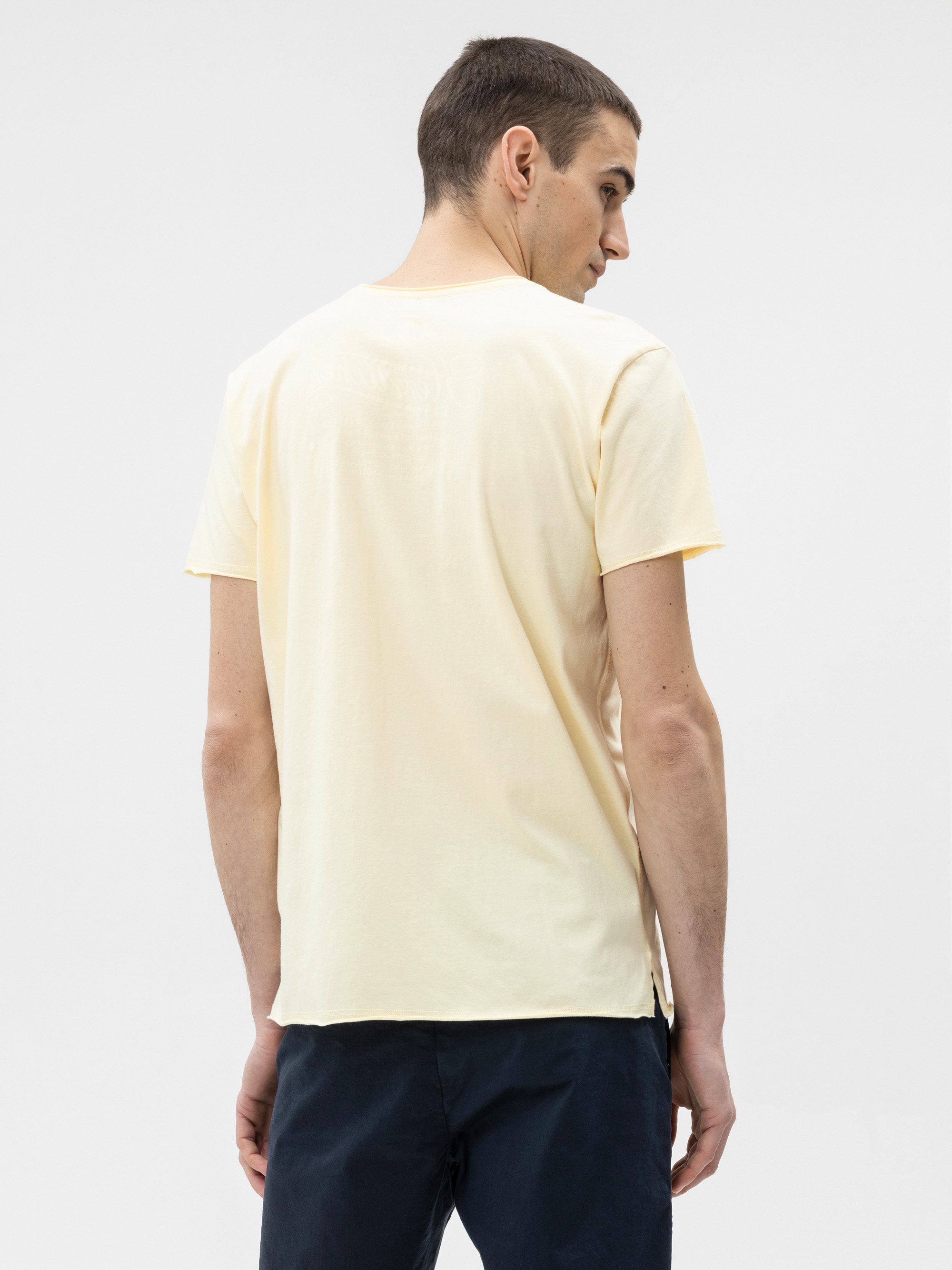 Ein Mann mit kurzen braunen Haaren steht mit dem Gesicht zur Wand und trägt ein hellgelbes kurzärmeliges T-Shirt und eine dunkle Hose vor einem schlichten weißen Hintergrund.