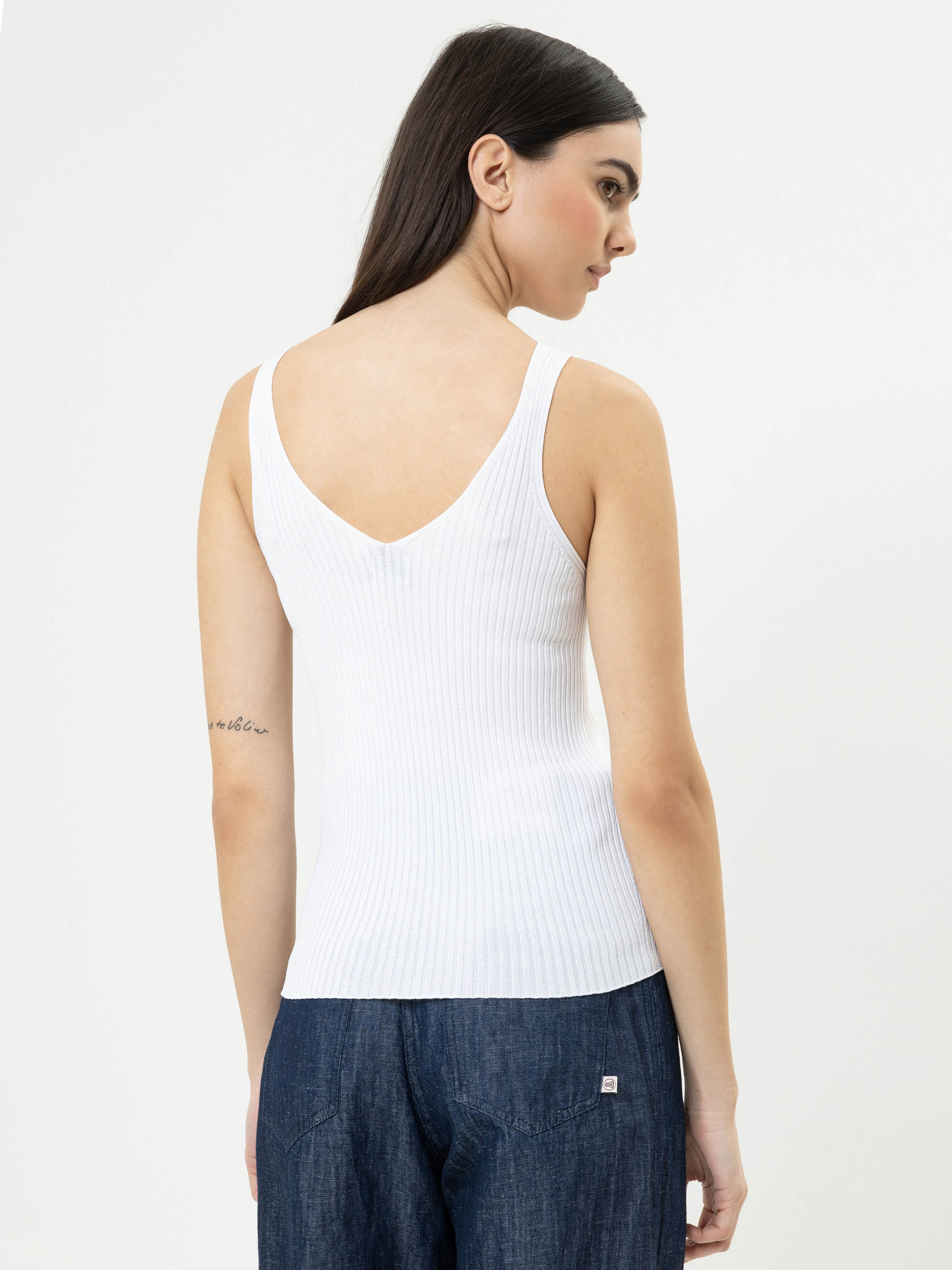 Eine Frau mit langen dunklen Haaren, bekleidet mit einem weißen, gerippten, ärmellosen Oberteil und blauen Jeans, steht mit dem Gesicht zur Wand und schaut über ihre Schulter. Auf ihrem linken Oberarm ist eine kleine Tätowierung zu sehen. Der Hintergr