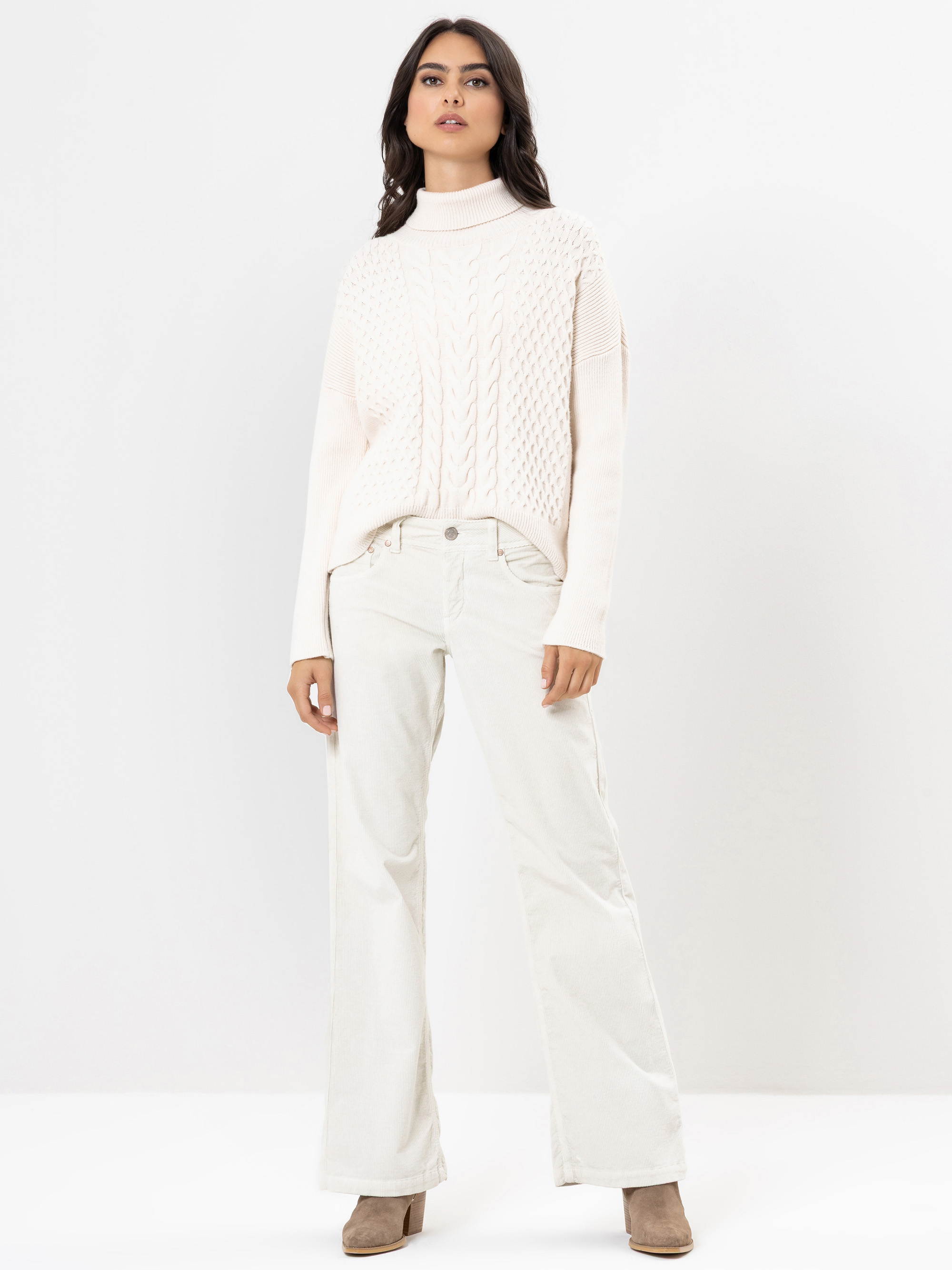 Eine Frau steht vor einem weißen Hintergrund und trägt einen elfenbeinfarbenen Rollkragenpullover mit Zopfmuster, eine elfenbeinfarbene Hose mit weitem Bein und beigefarbene hochhackige Stiefel. Sie blickt selbstbewusst in die Kamera und hat ihre Hände