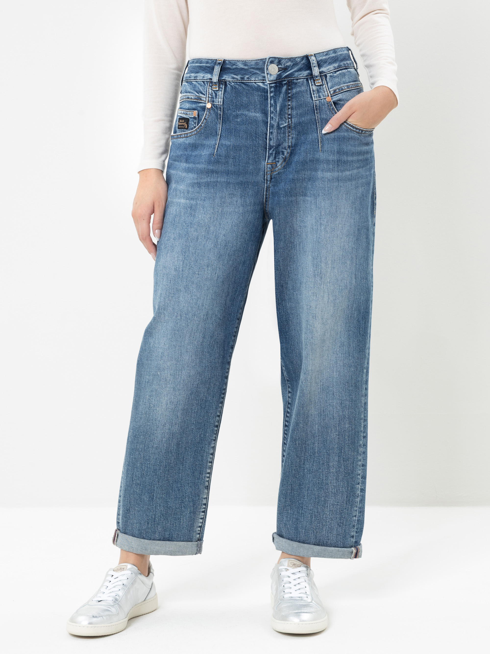 Eine Person, die eine blaue Jeans mit weitem Bein, ein weißes Langarm-Oberteil und silberne Sneaker trägt, steht mit einer Hand in der Tasche vor einem schlichten weißen Hintergrund. Die Jeans ist an den Knöcheln umgeschlagen.