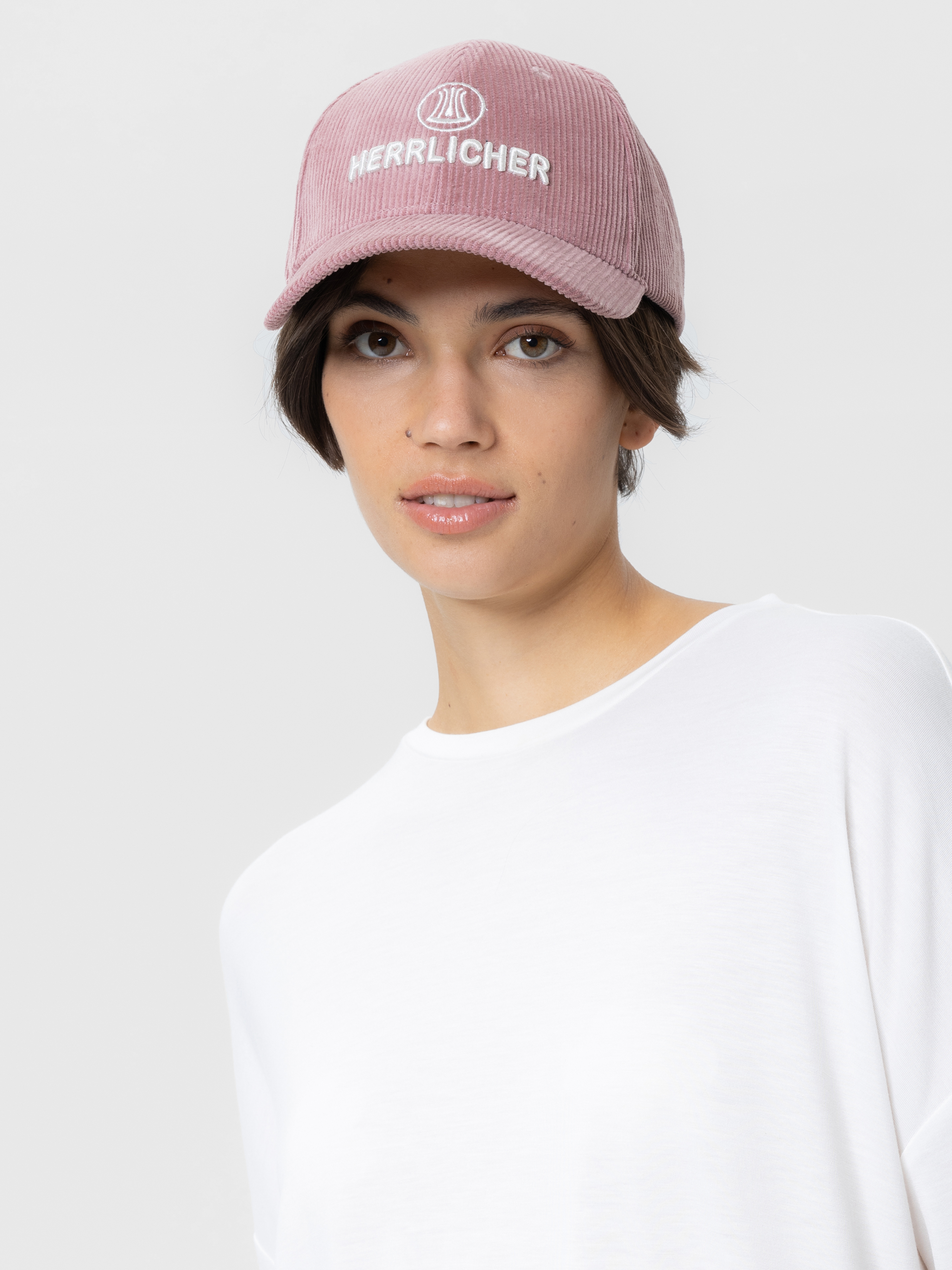 Eine Frau, die eine hellrosa Kordmütze mit dem aufgestickten Wort "Herrlicher" und ein schlichtes weißes Oberteil trägt, blickt leicht zur Seite vor einem schlichten hellen Hintergrund.
