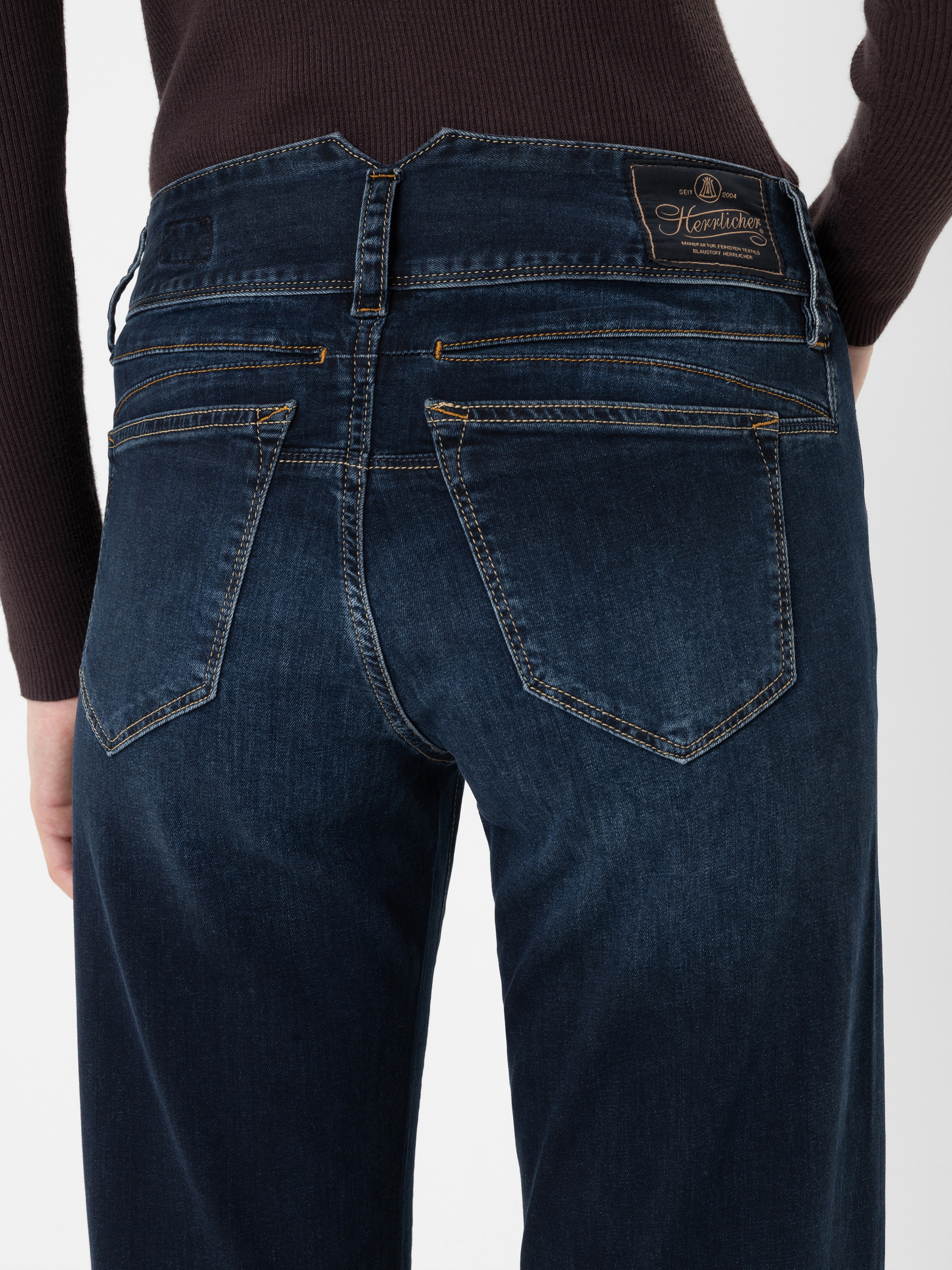 Eine Person, die dunkelblaue Jeans mit gelben Nähten trägt, von hinten gesehen. Die Jeans hat zwei Gesäßtaschen, zwei kleinere horizontale Taschen darüber und einen braunen Lederaufnäher am Bund. Die Person trägt außerdem einen dunklen Pullover.