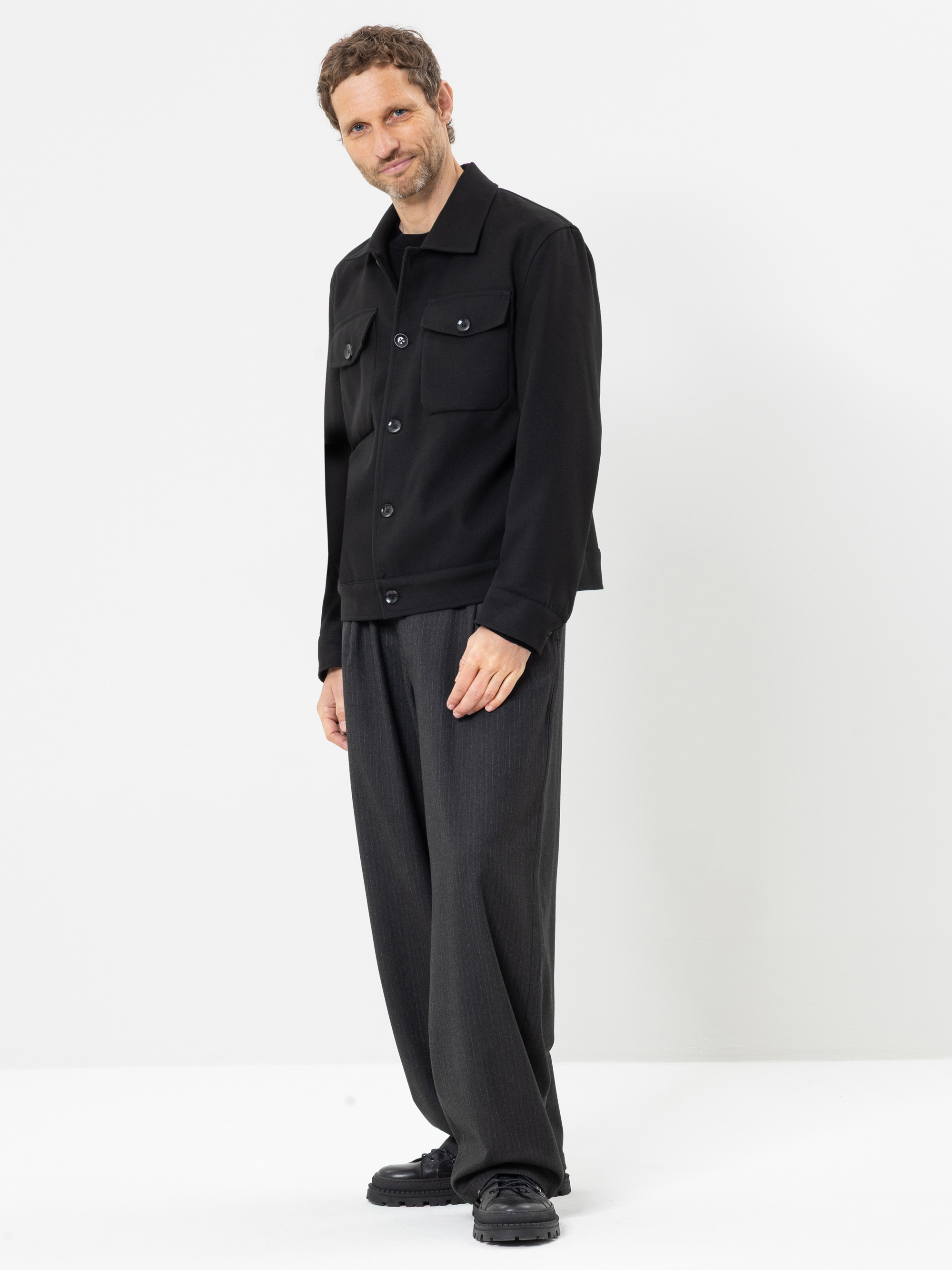Ein Mann steht vor einem weißen Hintergrund und lächelt. Er trägt eine schwarze Jacke mit Knöpfen über einem schwarzen Hemd, eine weite dunkelgraue Hose und schwarze Schuhe.