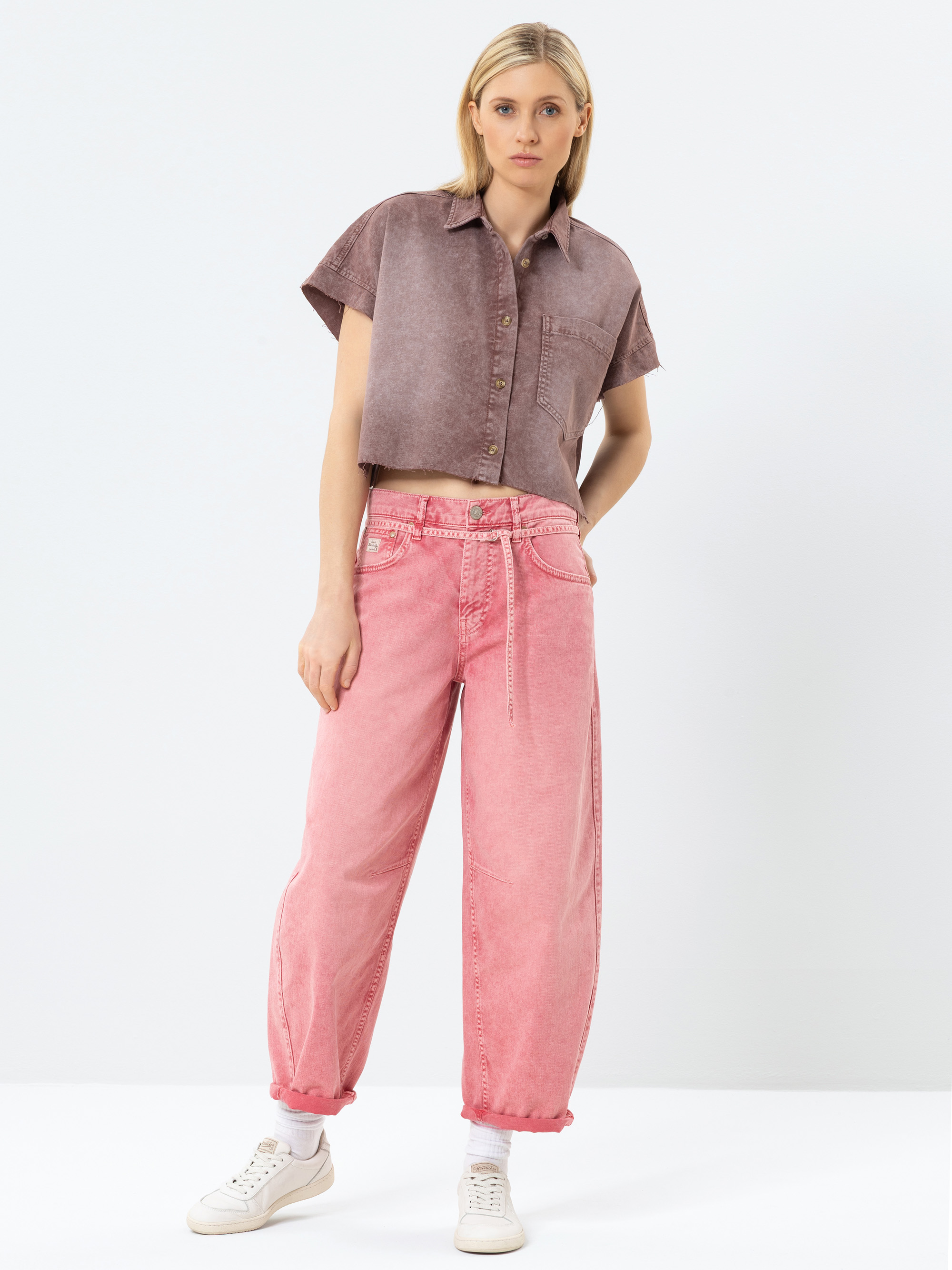 Eine Frau steht vor einem schlichten Hintergrund und trägt ein kurzärmeliges, geknöpftes, verblichenes lila Crop-Top und locker sitzende rosa Jeans mit gekrempelten Manschetten und einem Seilgürtel, dazu weiße Turnschuhe.