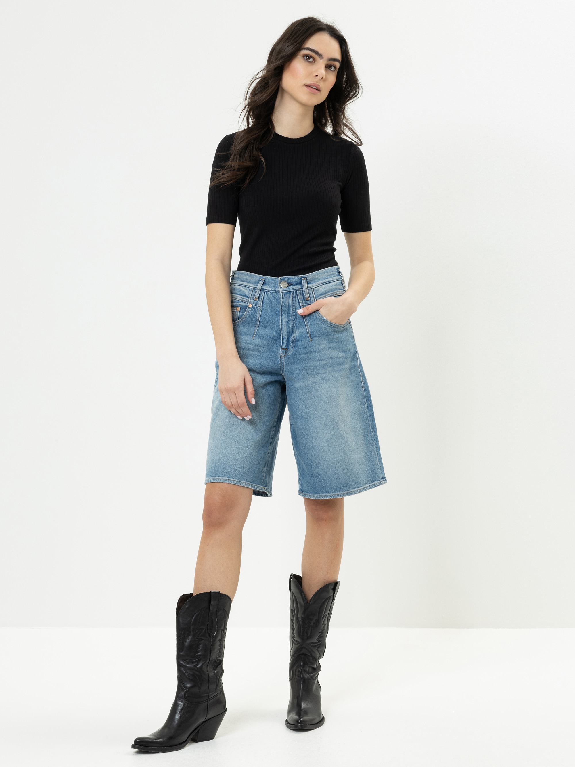 Eine Frau steht vor einem schlichten weißen Hintergrund und trägt ein schwarzes kurzärmeliges Oberteil, knielange blaue Jeans-Shorts und schwarze Cowboystiefel. Eine Hand steckt in ihrer Tasche, und sie hat langes, gewelltes dunkles Haar.