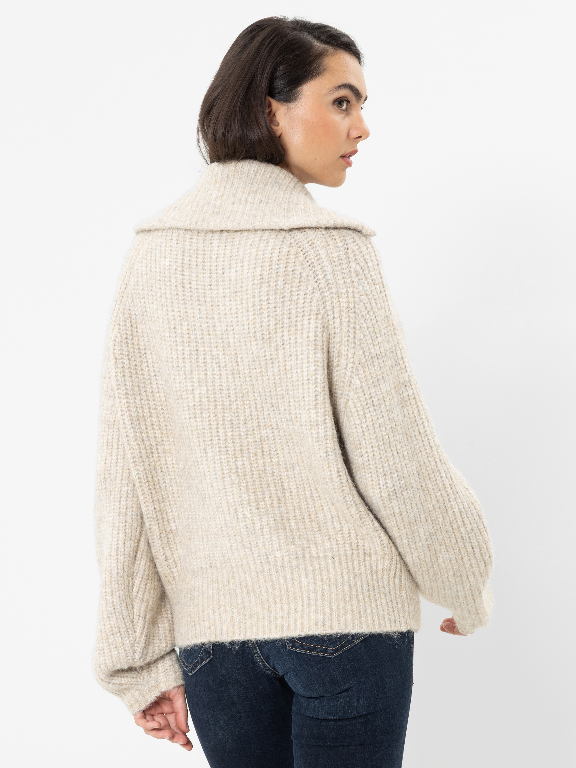 Eine Frau mit kurzen, dunklen Haaren trägt einen cremefarbenen, grob gestrickten Pullover mit weitem Kragen und dazu eine dunkelblaue Jeans. Sie steht vor einem schlichten weißen Hintergrund und blickt über ihre Schulter.