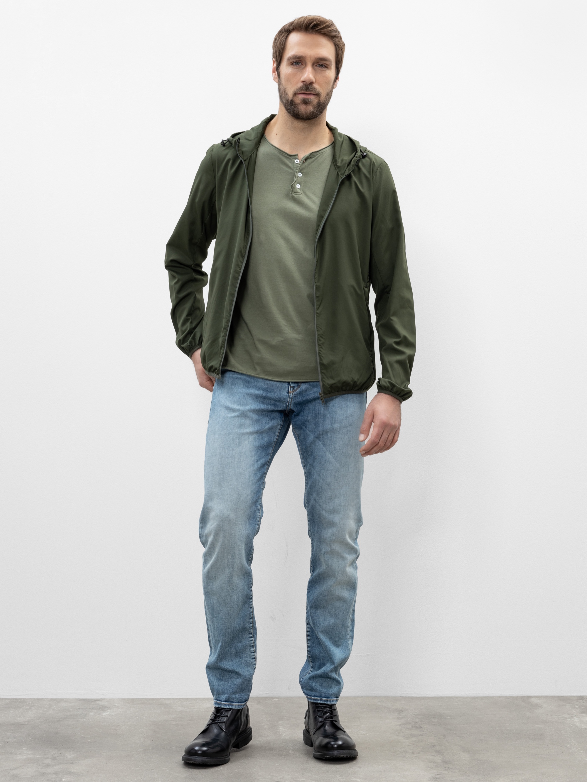 Ein Mann steht vor einer weißen Wand und trägt eine grüne Kapuzenjacke, ein passendes grünes Hemd, hellblaue Jeans und schwarze Stiefel. Er hat kurzes braunes Haar und einen Bart, eine Hand steckt in der Tasche seiner Jeans.
