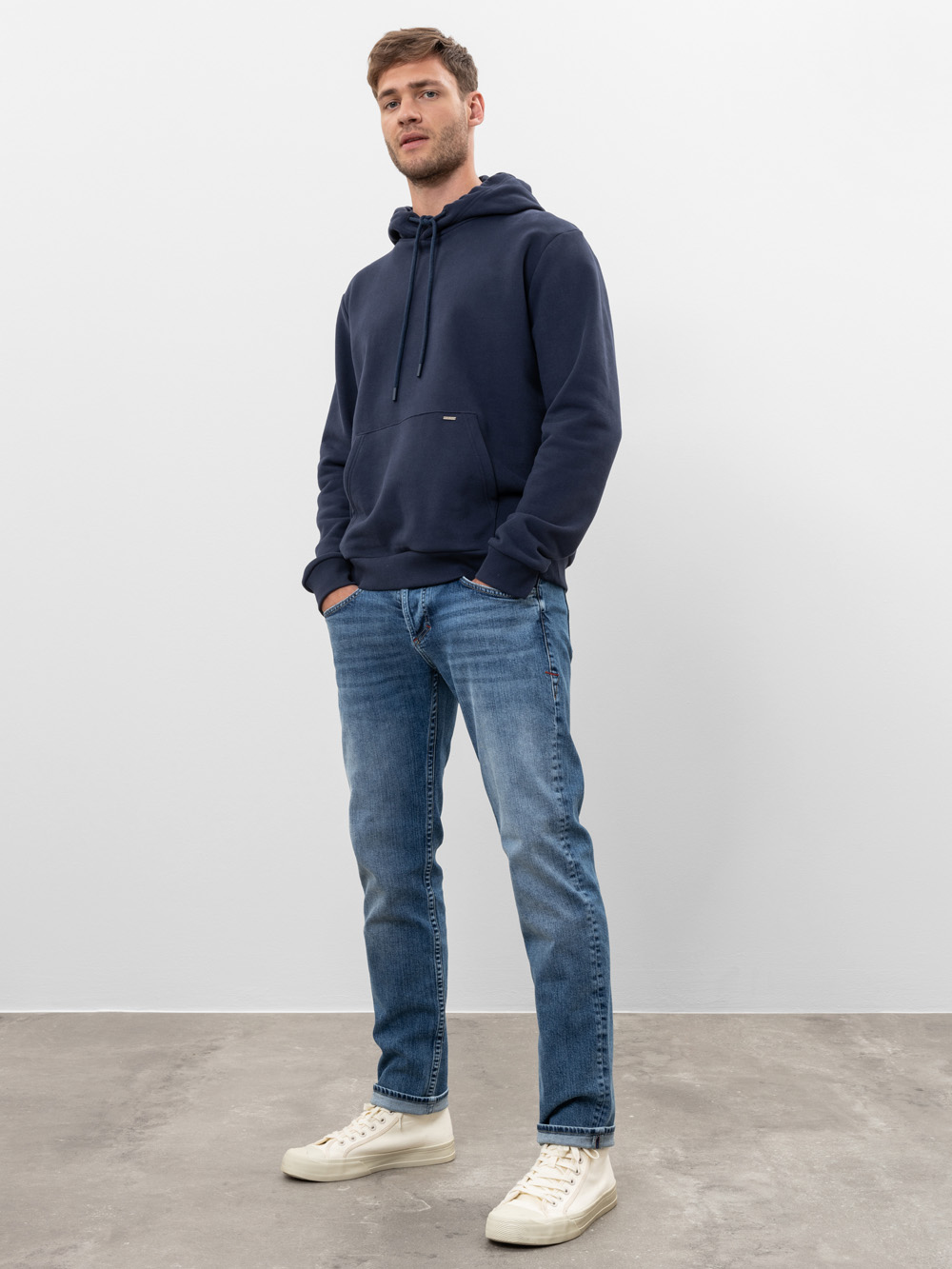 Ein Mann steht in einem Haus und trägt einen marineblauen Kapuzenpullover, blaue Jeans und weiße Turnschuhe. Er hat kurzes braunes Haar, seine Hände stecken in den Hosentaschen, und er steht vor einer schlichten weißen Wand auf einem Betonboden.
