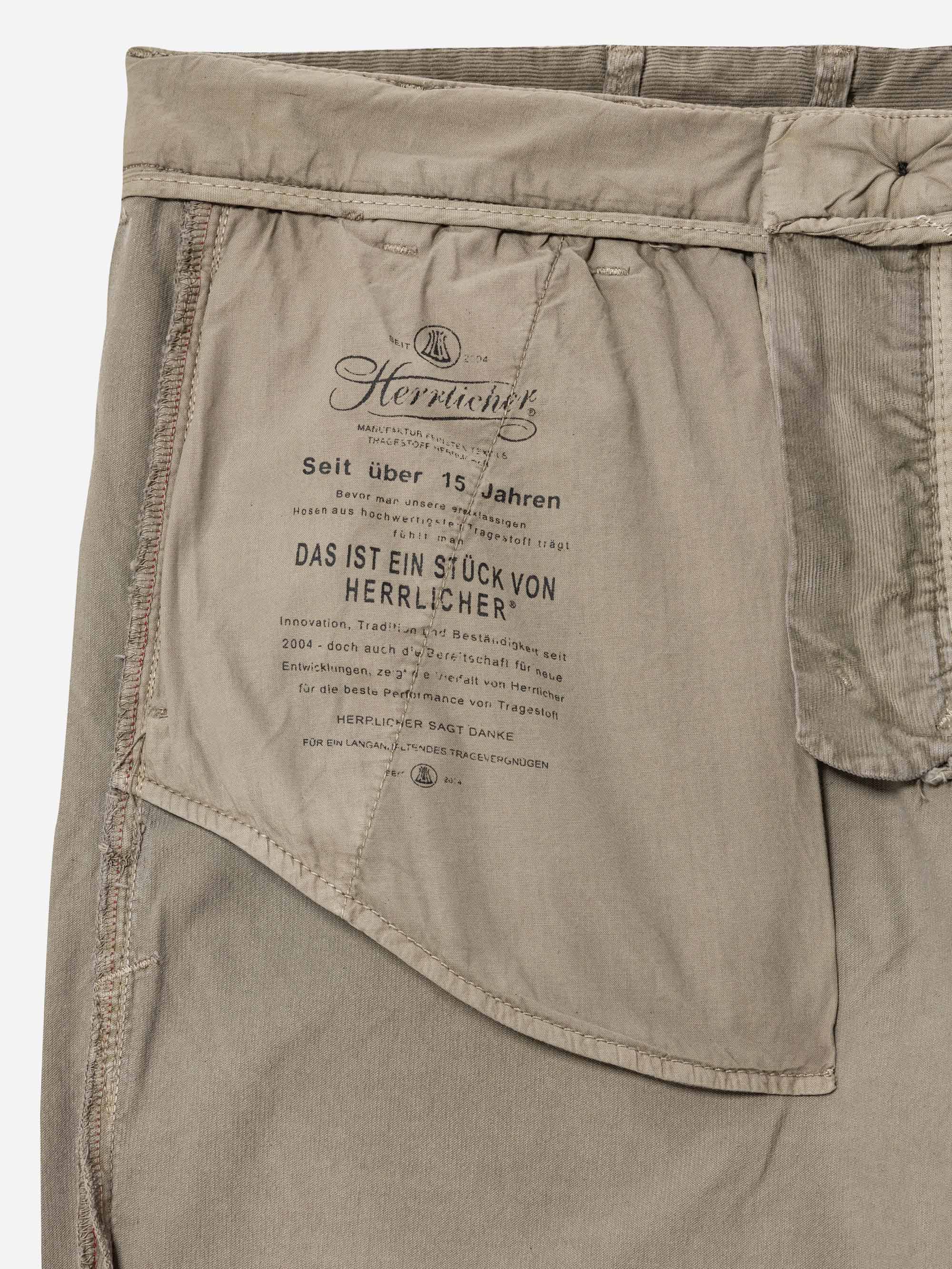 Nahaufnahme der Innentasche einer beigen Hose mit schwarzem Text, der in deutscher Sprache aufgedruckt ist, einschließlich Markenzeichen und Pflegeinformationen der Firma Herrlicher. Die Nähte und die Struktur des Stoffes sind deutlich sichtbar.