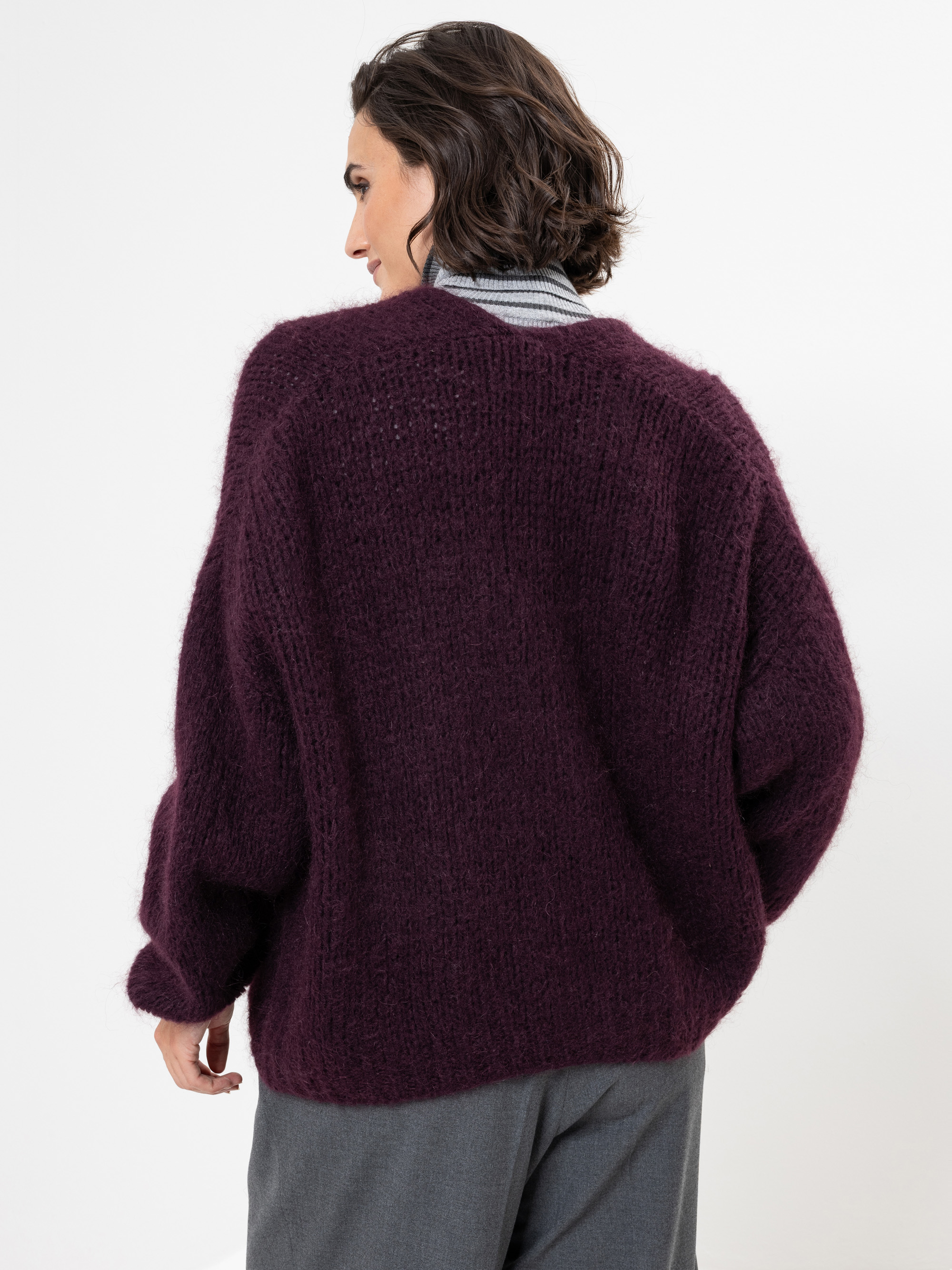 Eine Person mit kurzen dunklen Haaren, die einen lockeren, übergroßen, dunkelvioletten Strickpullover und eine graue Hose trägt, steht mit dem Gesicht von der Kamera weg vor einem schlichten weißen Hintergrund.