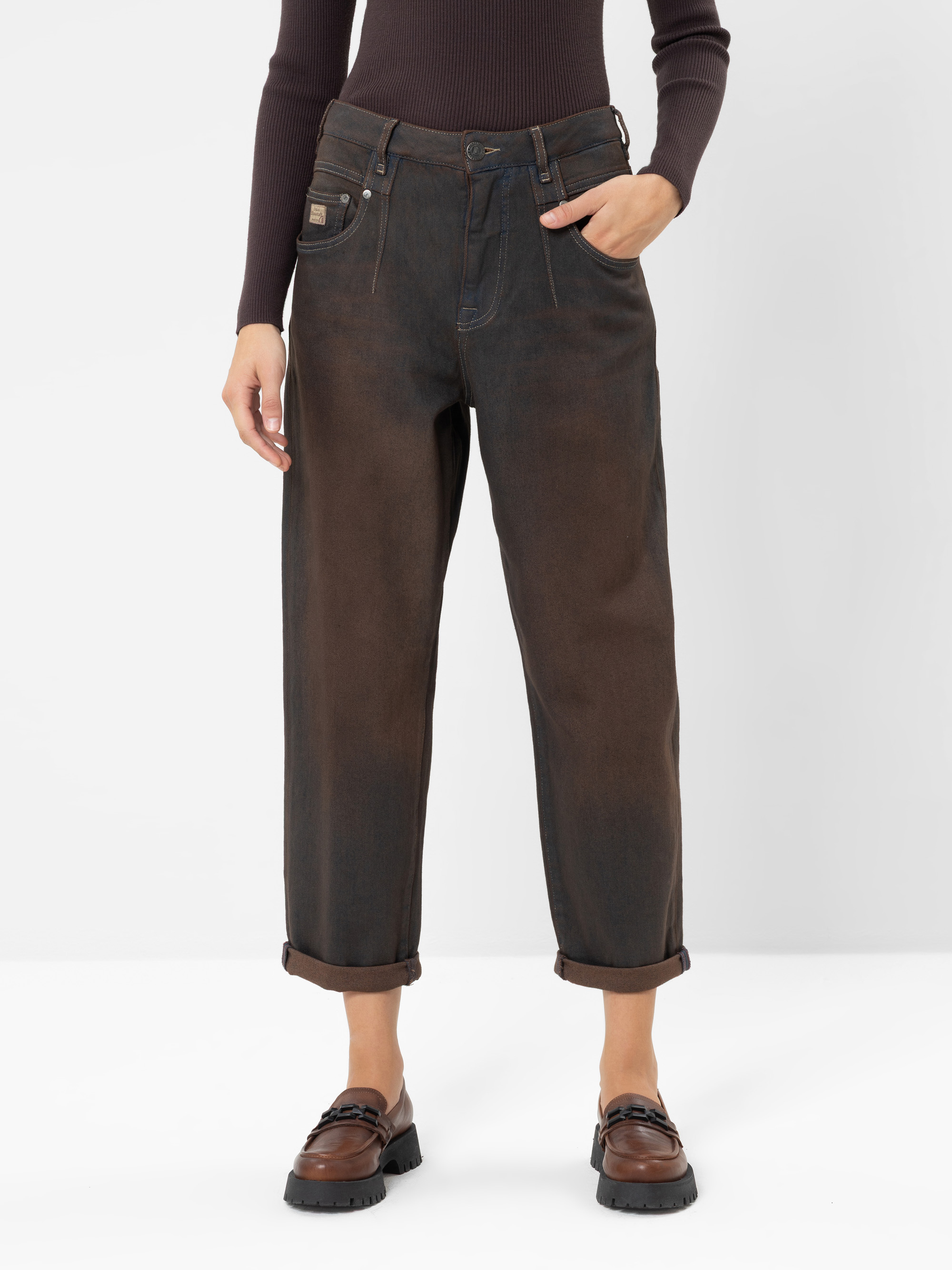 Eine Person, die eine braune, weit geschnittene Jeans mit Umschlag, ein eng anliegendes Langarm-Oberteil und braune Slipper trägt, steht vor einem schlichten weißen Hintergrund und hat eine Hand in der Tasche. Ihr Gesicht ist nicht zu sehen.