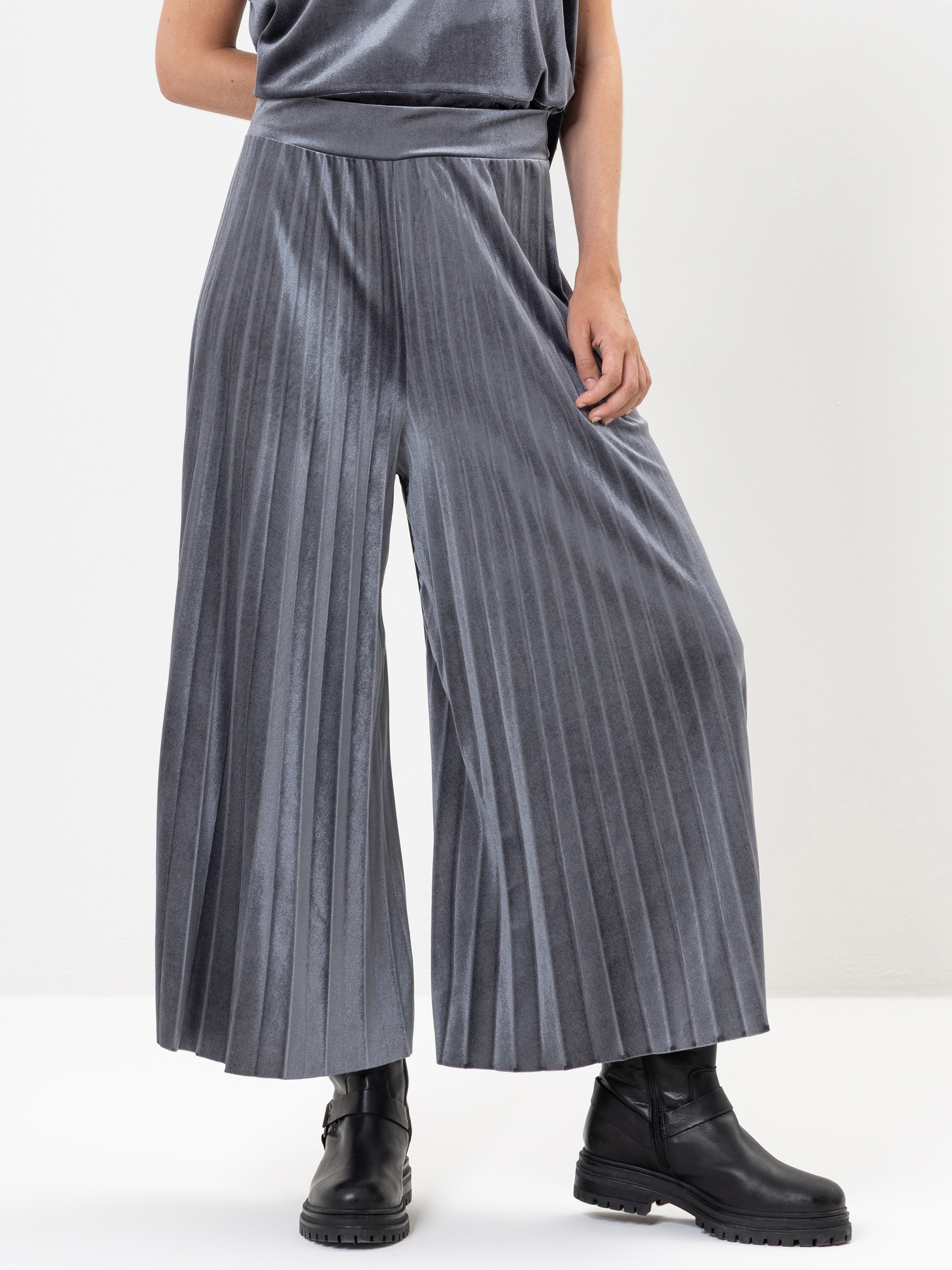 Eine Person, die eine weite, plissierte, metallgraue Hose und schwarze Stiefeletten trägt, steht mit einer Hand auf der Hüfte vor einem schlichten weißen Hintergrund.