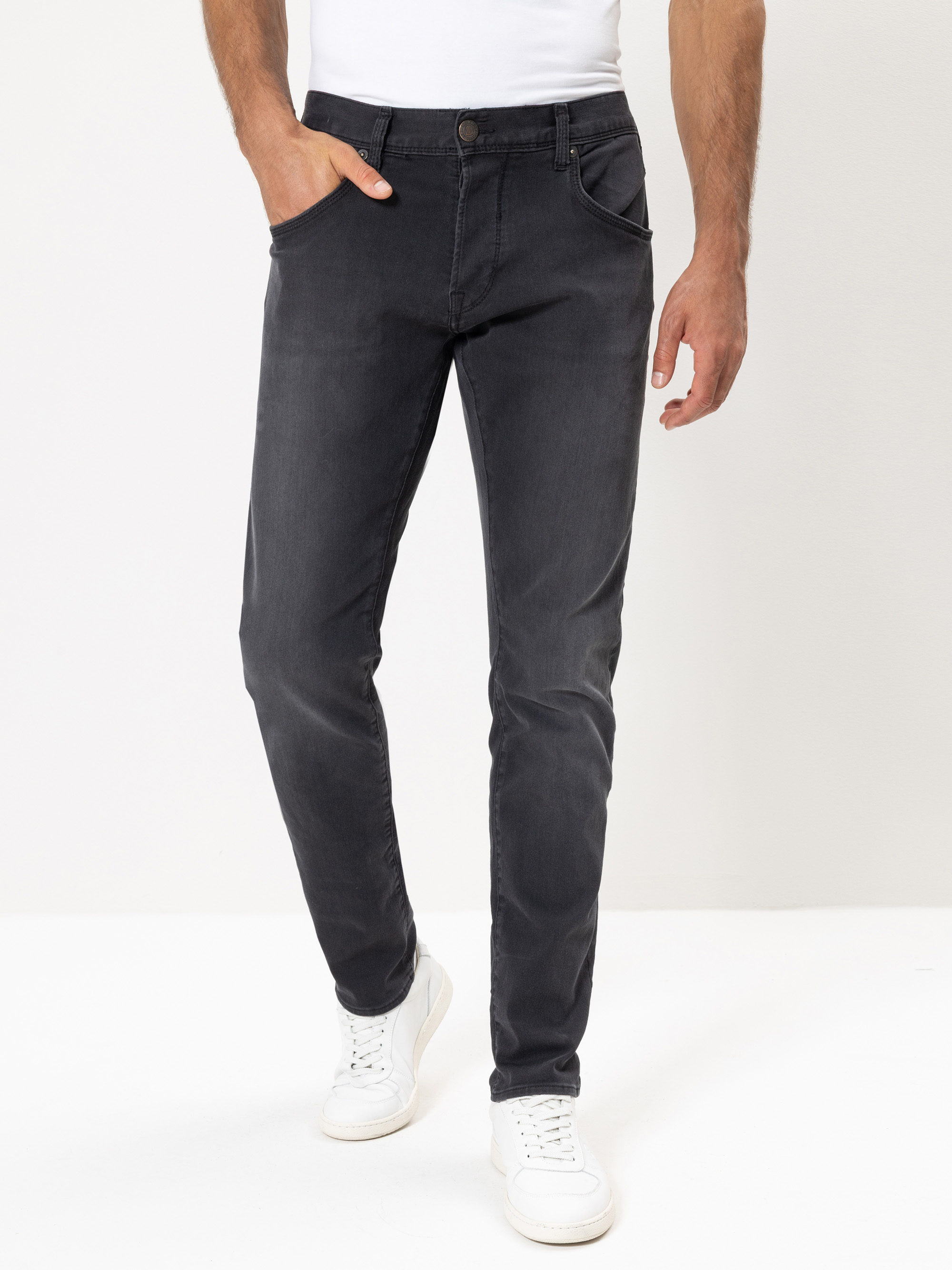 Ein Mann, der ein weißes T-Shirt, dunkelgraue Slim-Fit-Jeans und weiße Turnschuhe trägt, steht vor einem schlichten weißen Hintergrund und hat eine Hand in seiner Tasche.
