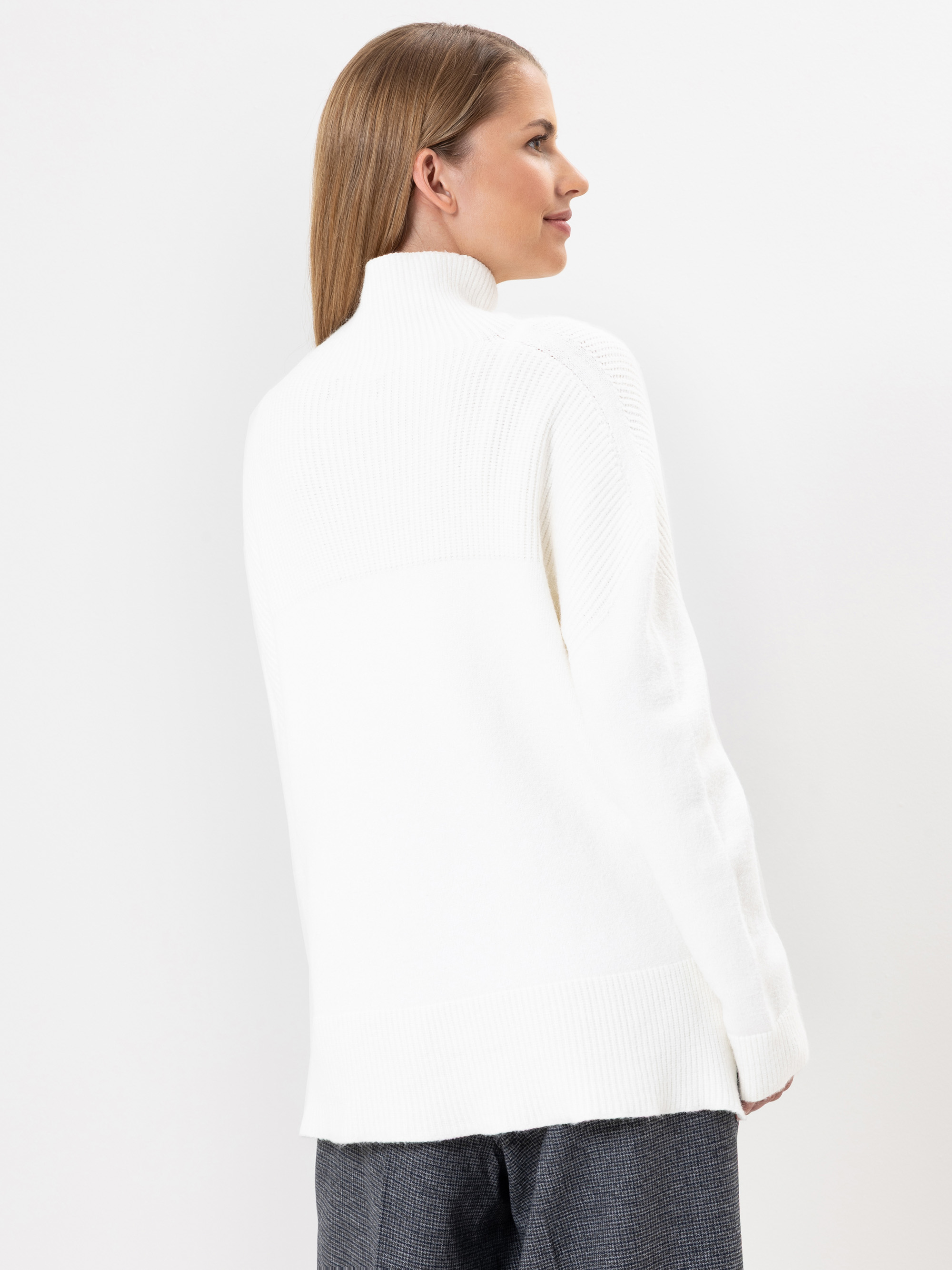 Eine Frau mit glattem, hellbraunem Haar trägt einen weißen Rollkragenpullover und eine graue Hose. Sie steht seitlich und blickt nach rechts vor einem schlichten weißen Hintergrund.