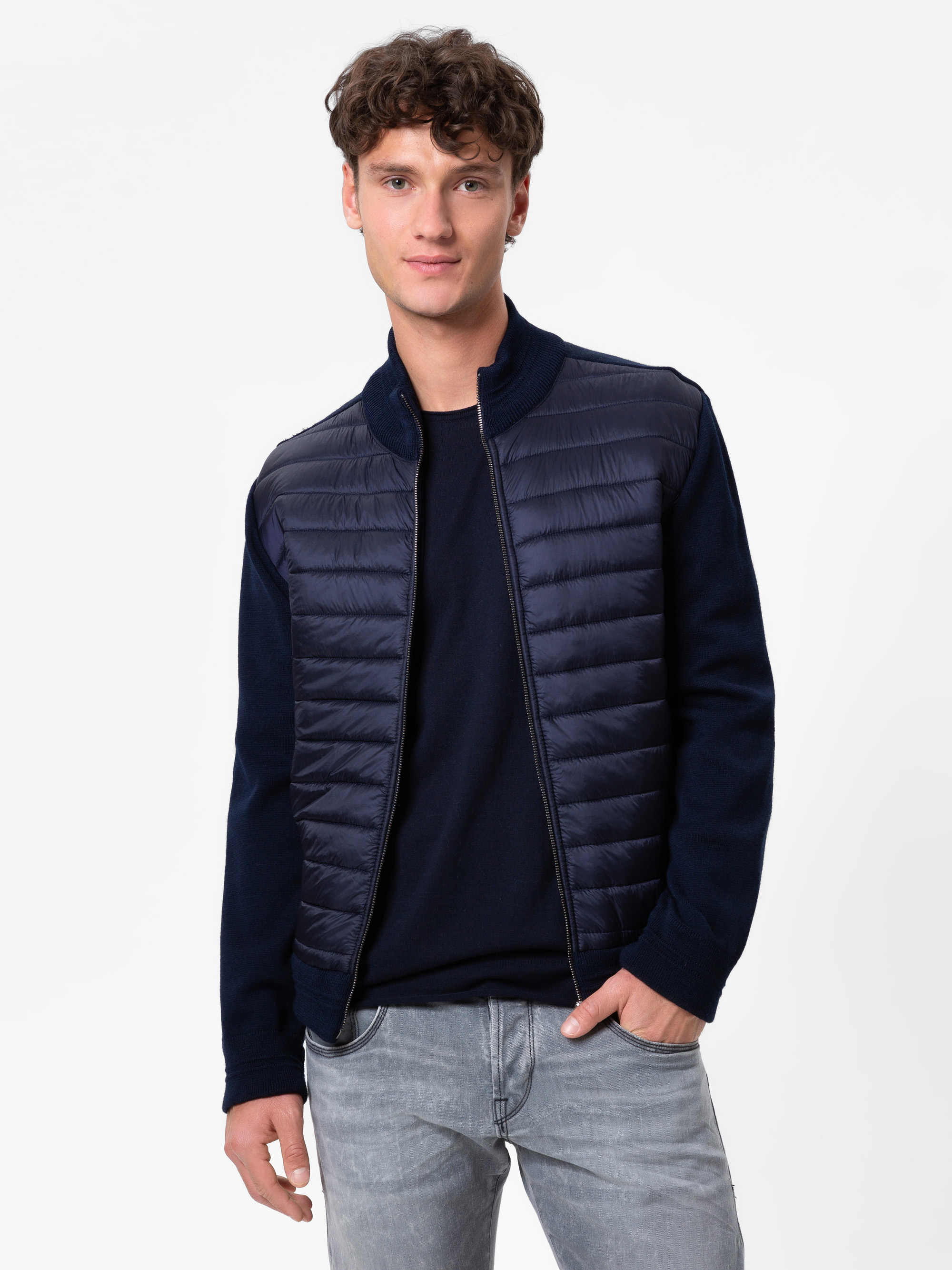Ein junger Mann mit lockigem braunem Haar trägt eine marineblaue Steppjacke über einem dunklen Hemd und hellgraue Jeans und steht mit neutralem Gesichtsausdruck vor einem schlichten weißen Hintergrund.