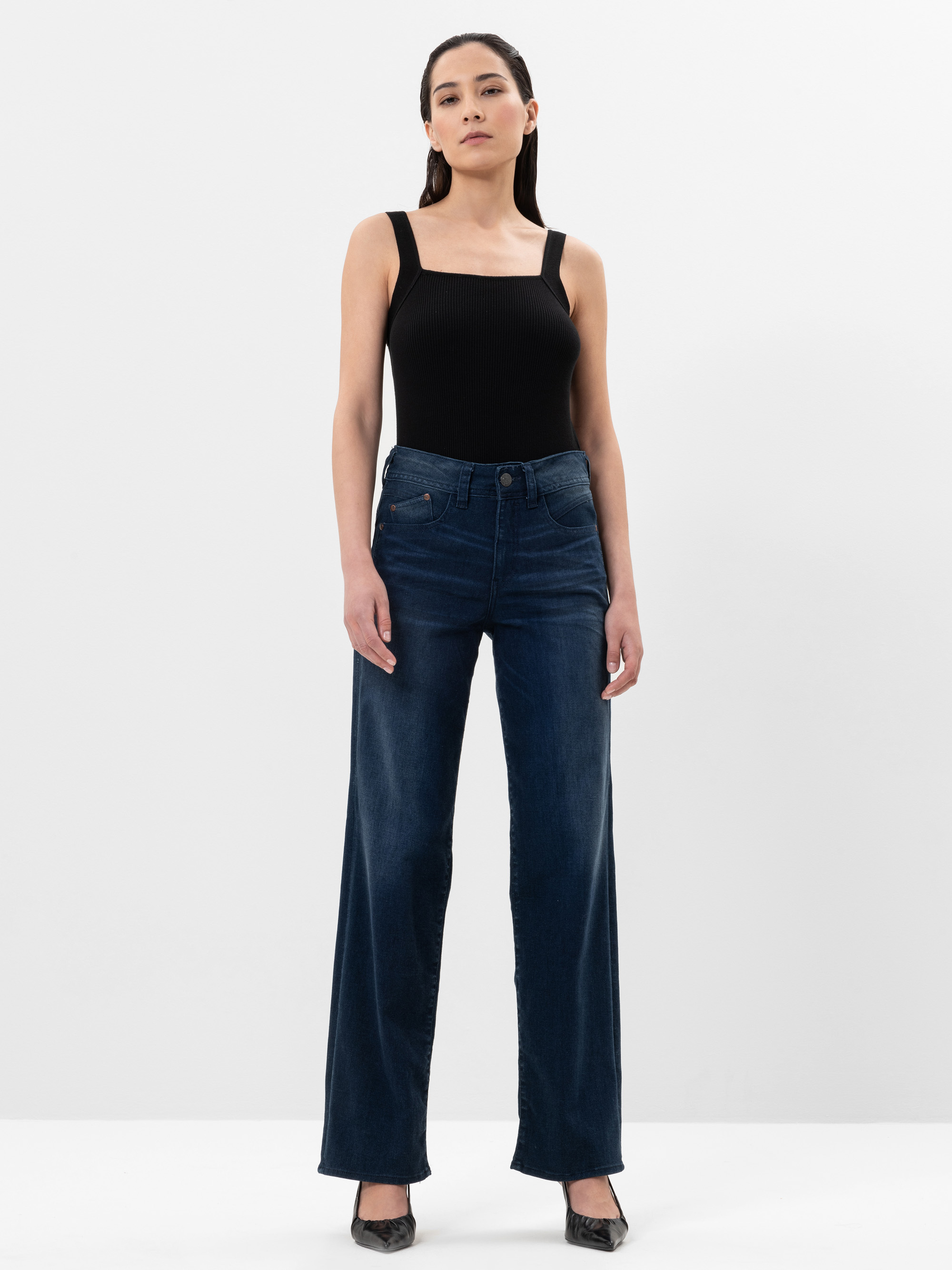 Eine Frau steht vor einem schlichten weißen Hintergrund und trägt ein schwarzes ärmelloses Oberteil mit quadratischem Ausschnitt, dunkelblaue Jeans mit hoher Taille und weiten Beinen sowie schwarze Schuhe mit spitzen Zehen. Sie hat glattes, dunkles Haa