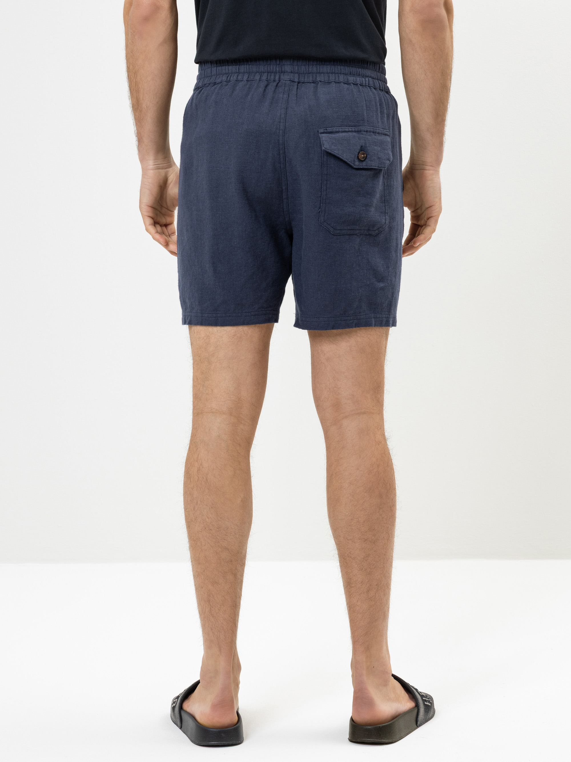 Eine Person, die mit dem Rücken zur Kamera steht und ein schwarzes T-Shirt, marineblaue Shorts mit Gesäßtasche und Knopf sowie schwarze Gleitsandalen vor einem schlichten weißen Hintergrund trägt.