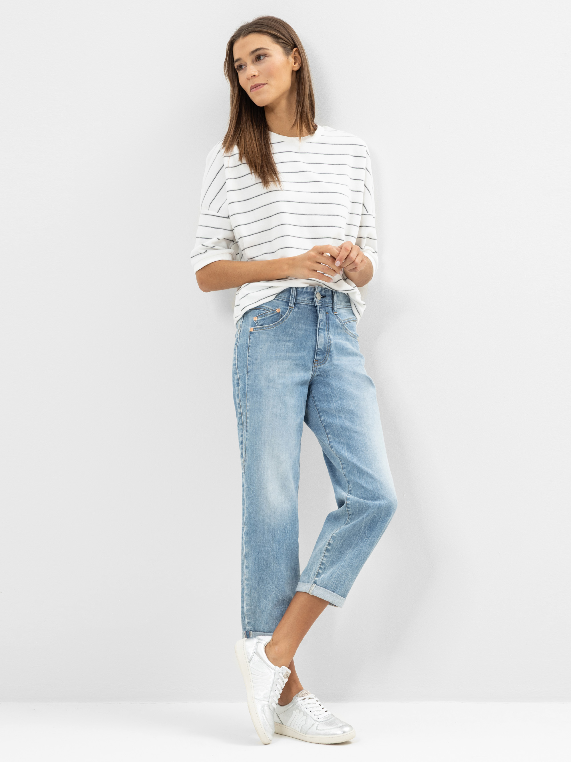 Eine Frau steht vor einer weißen Wand und trägt ein weißes, kurzärmeliges, gestreiftes Hemd, hellblaue Jeans mit Bündchen und weiße Turnschuhe. Sie hat langes, glattes Haar und blickt mit verschränkten Händen zur Seite.