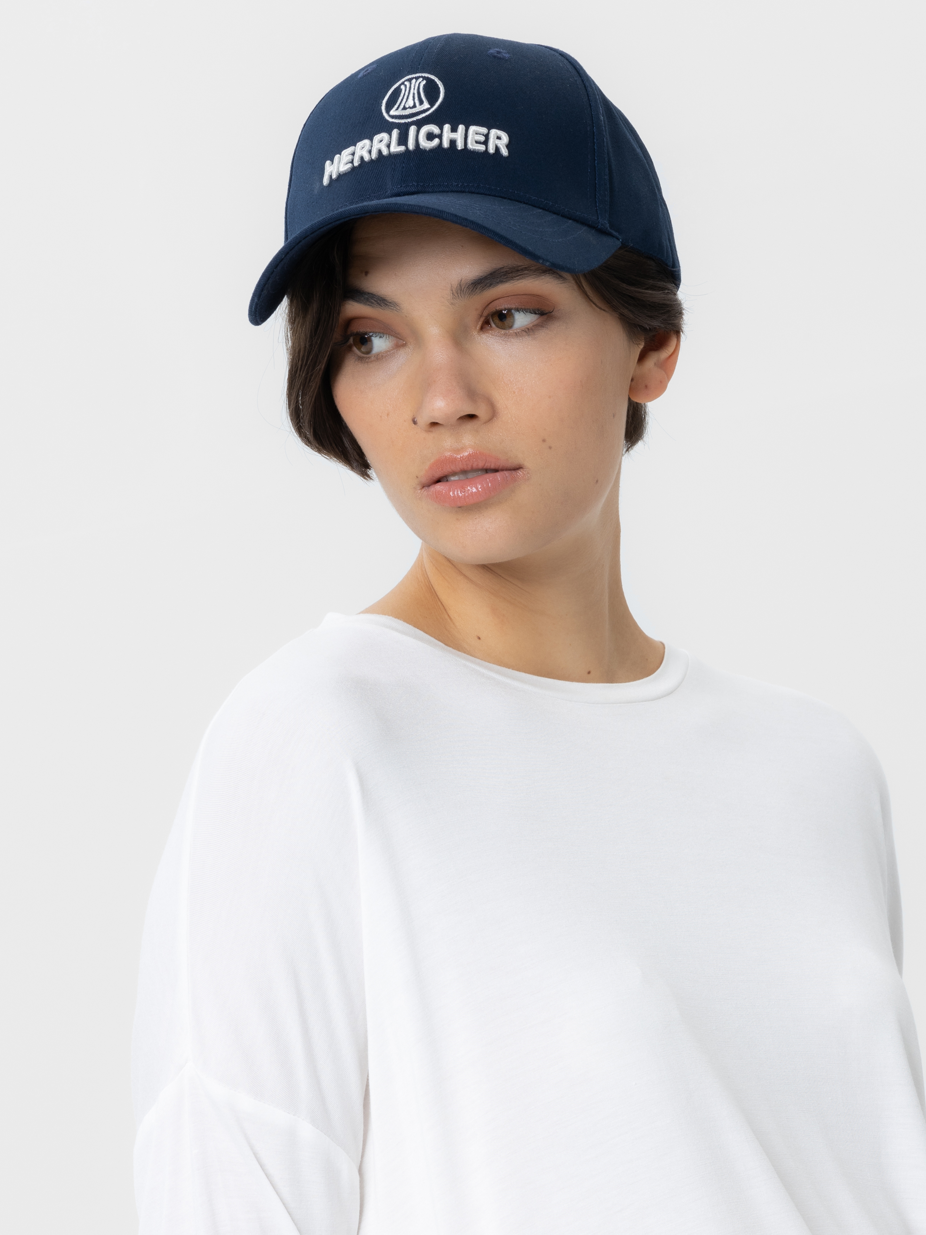 Eine Person, die eine marineblaue Baseballmütze mit der Aufschrift HERLICHER in weißer Schrift und ein weißes Oberteil trägt, schaut vor einem schlichten hellen Hintergrund zur Seite.