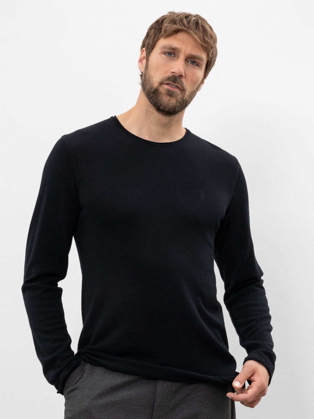 Ein Mann mit kurzen braunen Haaren und einem Bart trägt einen schwarzen, langärmeligen Pullover und eine graue Hose und steht vor einem schlichten weißen Hintergrund. Er blickt direkt in die Kamera und hat einen neutralen Blick.